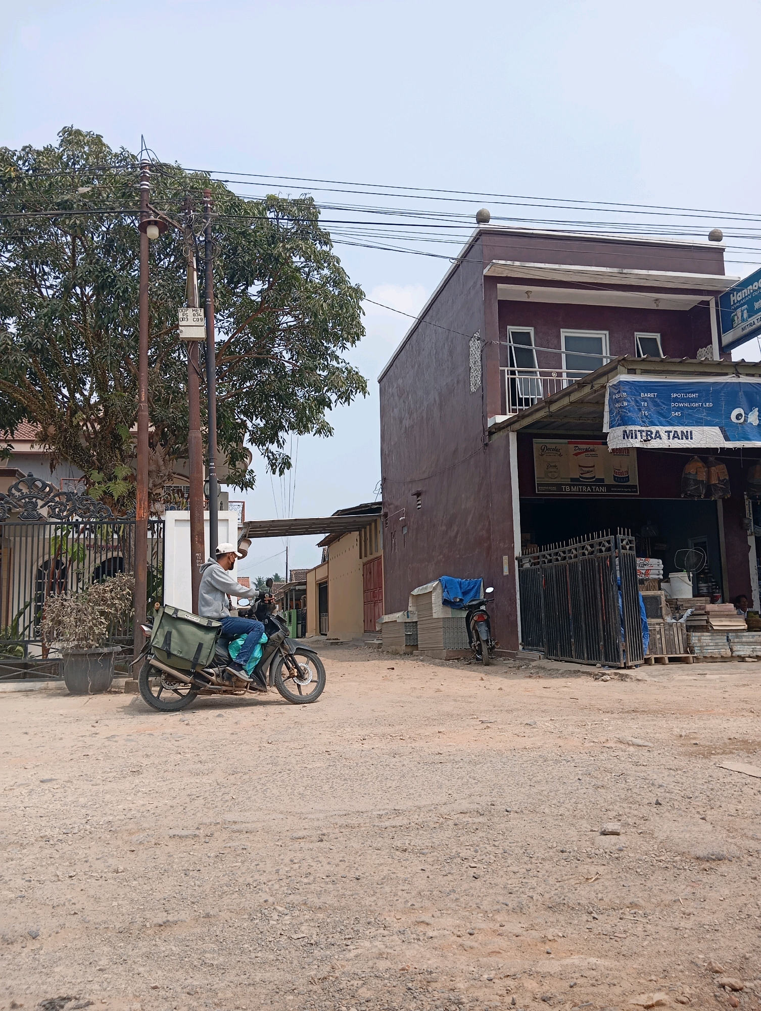 In the city of Leman Benawi, Indonesia, a man wearing a grey hoodie, blue jeans, and a white cap is riding a motorcycle with a green saddlebag on the back. The motorcycle is traveling on a dirt road. To the right of the road is a two-story building with a maroon facade and a business called "Mitra Tani" on the first floor, which appears to sell building materials and agricultural supplies. The building has a balcony on the second floor and several windows. To the left of the road is a large tree and a black metal fence. The sky is overcast.