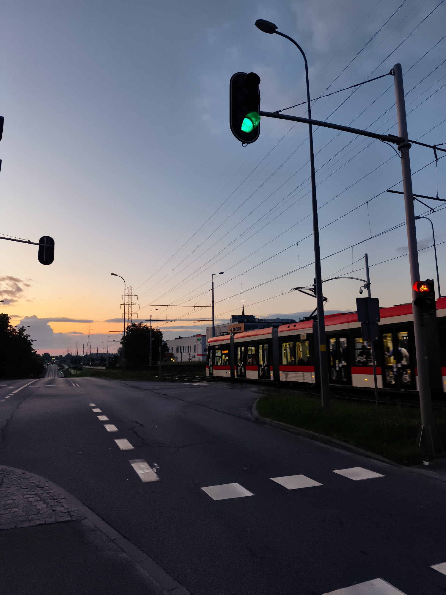 The image shows a street scene in Gdańsk, Poland, during twilight. A tram with red and white markings is moving on the right side of the frame. Traffic lights are visible, with one showing a green signal and another showing a red signal with a bicycle icon. The road has white markings and a pedestrian crossing. Power lines stretch across the sky, supported by poles. The sky displays a gradient of colors from the setting sun, with some clouds present. The overall scene depicts an urban environment at dusk, with active transportation.