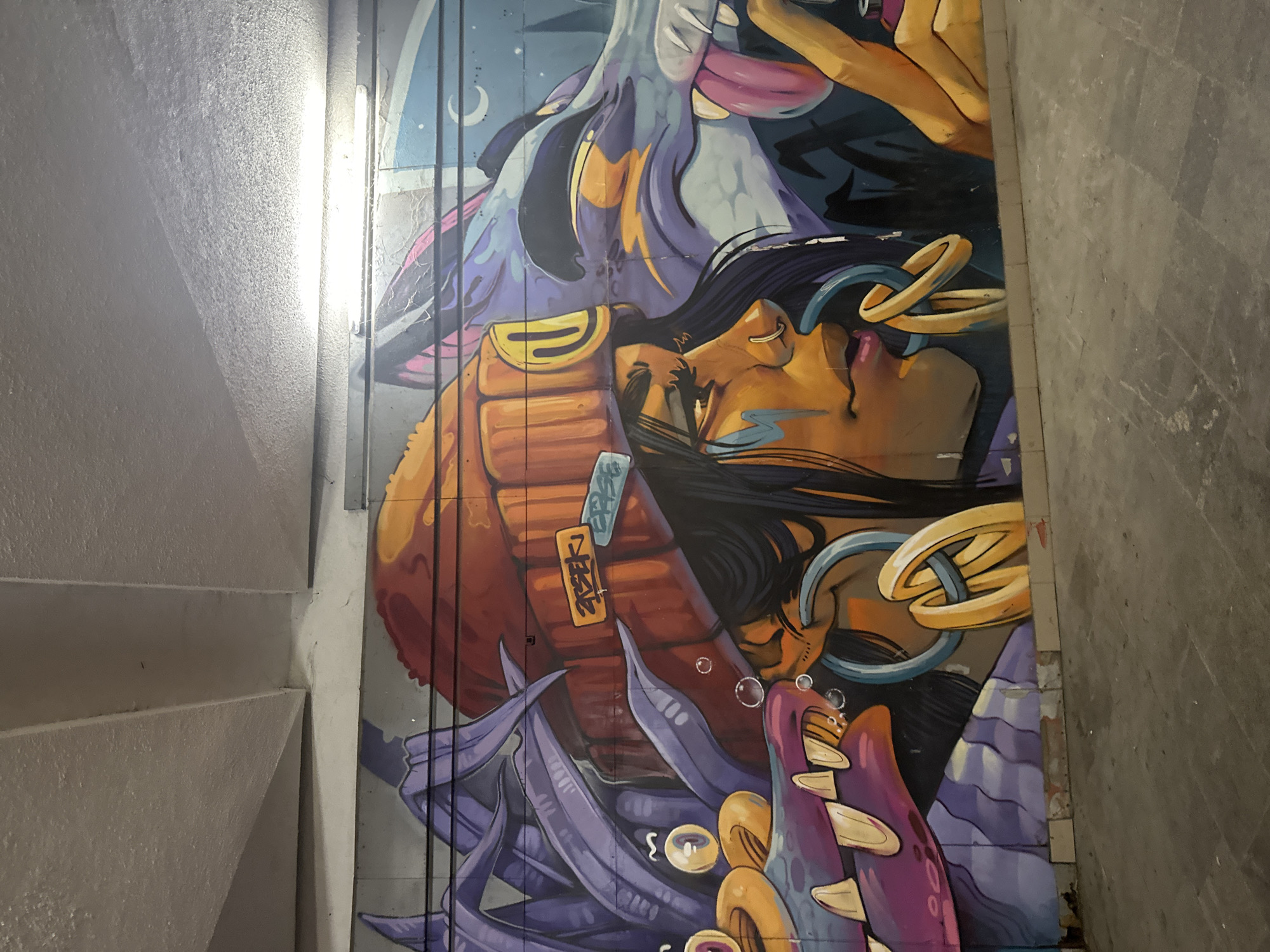 A striking mural adorns a wall in Sofia, Bulgaria. The mural features a person with dark hair and golden skin tones with what appears to be the face of a crocodile. The figure is adorned with golden rings and surrounded by what appears to be various creatures and plant life. The color palette is rich and vibrant, incorporating shades of purple, gold, and orange. The surrounding environment consists of a concrete wall and what appears to be stairs. The image is taken from an elevated perspective, capturing the mural in its entirety.
