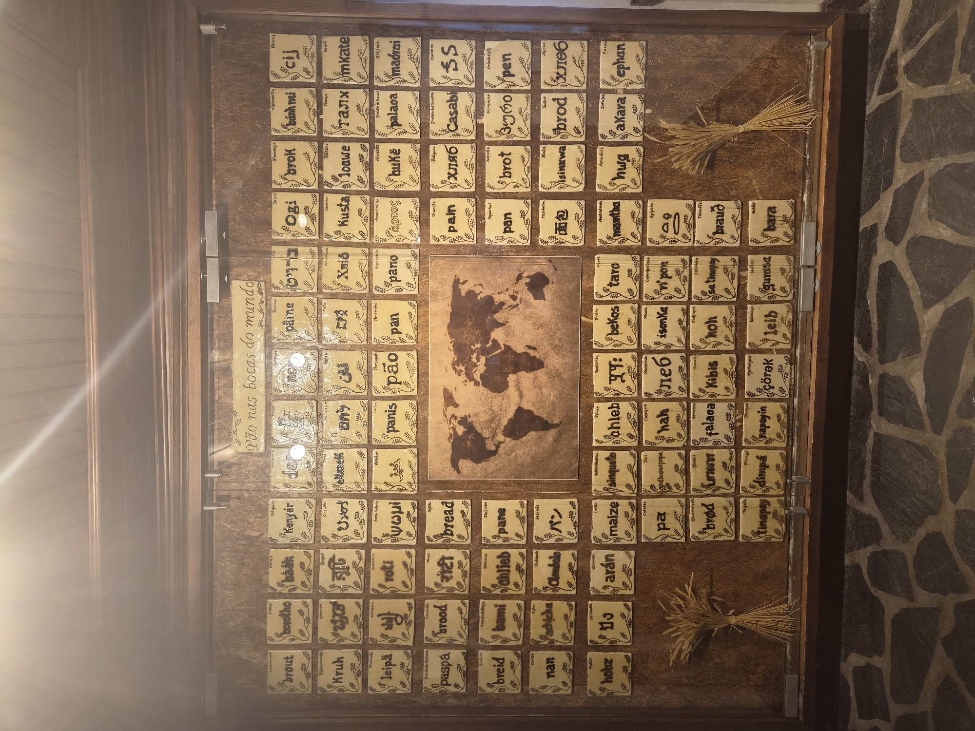 The image displays a wall-mounted exhibit featuring a world map and numerous small, square ceramic tiles, each adorned with a word written in a different language, likely representing the word "bread." The exhibit is located in Seia, Portugal, at the Museu do Pão (Bread Museum). The tiles are arranged in a grid-like pattern above and to the sides of the map. The overall backdrop of the exhibit appears to be a textured, wood-like material. The lighting suggests an indoor environment, possibly dimly lit to highlight the display. The bottom of the image shows a stone floor.