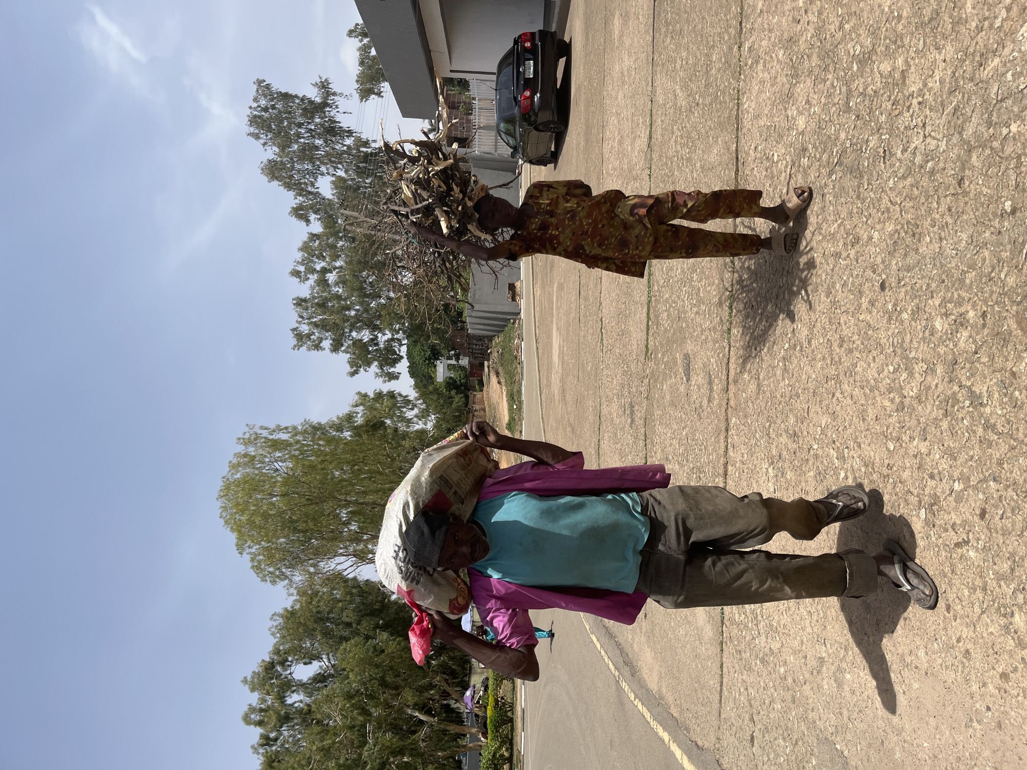 Two individuals are walking along a paved surface, likely a road or walkway, in what appears to be an outdoor setting. The first individual, closer to the foreground, carries a large sack or bundle over their shoulder, supported by their head. They are wearing a blue shirt, grey pants, a pink shawl draped over their shoulder, and sandals. The second individual, further in the background, carries a collection of sticks or branches atop their head. They are wearing a camouflage-patterned outfit and sandals. In the background, there are trees, buildings, and a dark-colored car. The sky is blue with some scattered clouds. Considering the prompt, the location is possibly within the city of Jos, Nigeria.