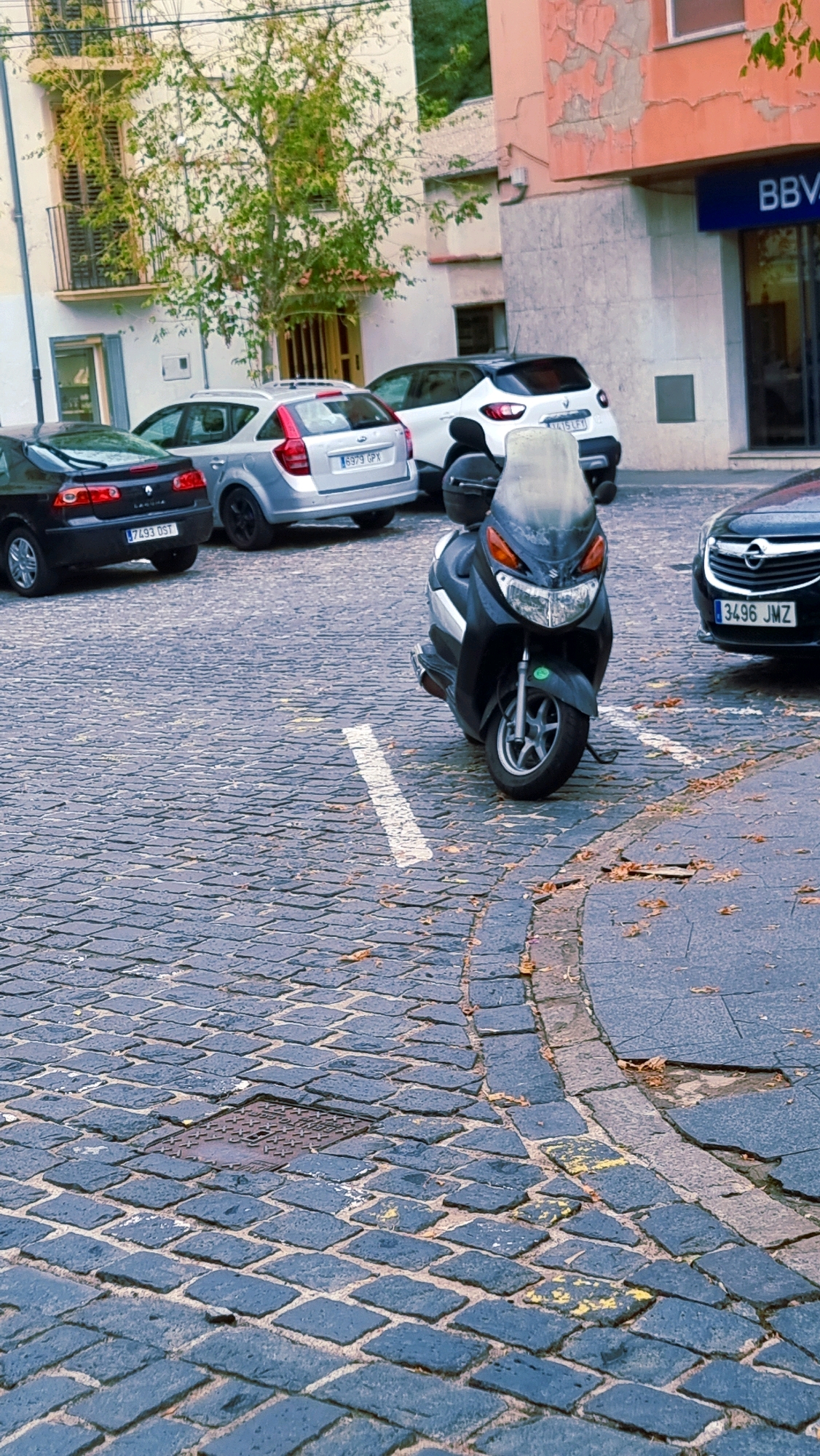 The image shows a street scene in Sant Joan Les Fonts, Spain. A black scooter is parked on a cobblestone street, facing forward. Behind the scooter, several cars are parked, including a silver hatchback and a white SUV. A black sedan is visible on the right side of the image. The buildings in the background appear to be a mix of residential and commercial, with one building featuring peeling paint and another with a blue BBVA bank sign. A tree with sparse foliage grows in front of one of the buildings. The ground is paved with dark cobblestones, with some fallen leaves scattered about. There are no people visible in the image.