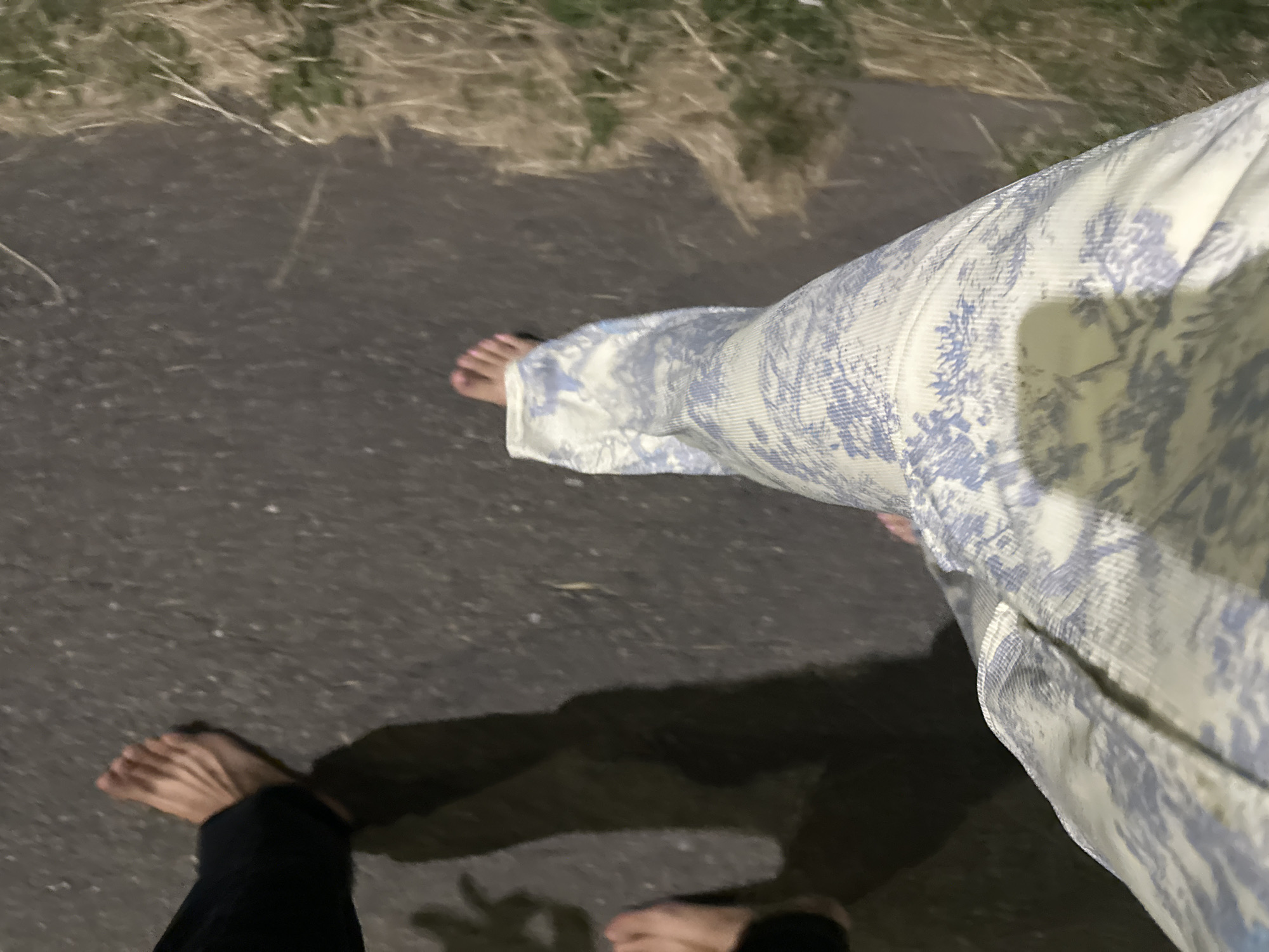 An overhead shot shows two pairs of feet walking on a paved surface, possibly a sidewalk or path, with dry grass on the right side of the frame. The left foot, belonging to someone wearing black pants, is bare. The right foot, belonging to someone wearing patterned pants, is also bare. The scene is dimly lit, suggesting it is either dusk or night. The location of Nevinnomyssk, Russia, is not discernible from the image itself.