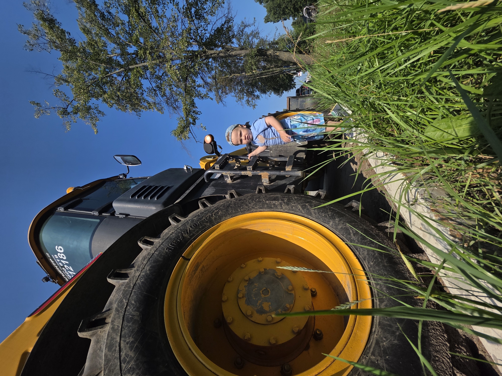 A low-angle, full-shot photograph captures a young boy standing next to a large yellow and black construction vehicle on a bright, sunny day. The boy, wearing a blue t-shirt, blue and white patterned shorts, and a patterned blue bandana, is looking towards the camera. He is positioned to the right of the vehicle's large tire, with his right hand resting on a metal step or railing. The construction vehicle, likely a backhoe loader, is dominant in the frame, with its massive tire and yellow rim in the foreground on the left. The cab of the vehicle is visible, displaying a blue tinted window with some text and numbers. The scene appears to be an outdoor location, possibly a construction site or an area adjacent to one, given the presence of the heavy machinery and the clear blue sky. Lush green grass and weeds grow along the edge of a paved or dirt surface where the vehicle is parked. In the background, a tall tree with green foliage stands against the sky, and other vehicles and structures are faintly visible, suggesting an urban or semi-urban environment. The photograph's perspective emphasizes the scale of the construction vehicle in relation to the child. There is no evidence to suggest this scene is specifically located in the city of Chaikovsky, Russia.