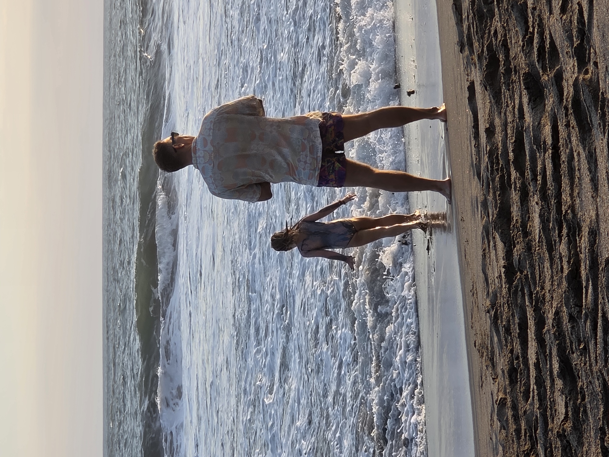 A full-shot, eye-level, vertical, outdoor, daytime, medium shot shows a man and a child on a beach in North Kuta, Indonesia. The man is facing away from the camera, looking at the ocean. He is wearing a short-sleeved patterned shirt and shorts. The child is also facing away from the camera, standing closer to the water, wearing a one-piece swimsuit. The ocean has waves and foam. The sand in the foreground is textured with tire tracks. The sky is a pale gradient.