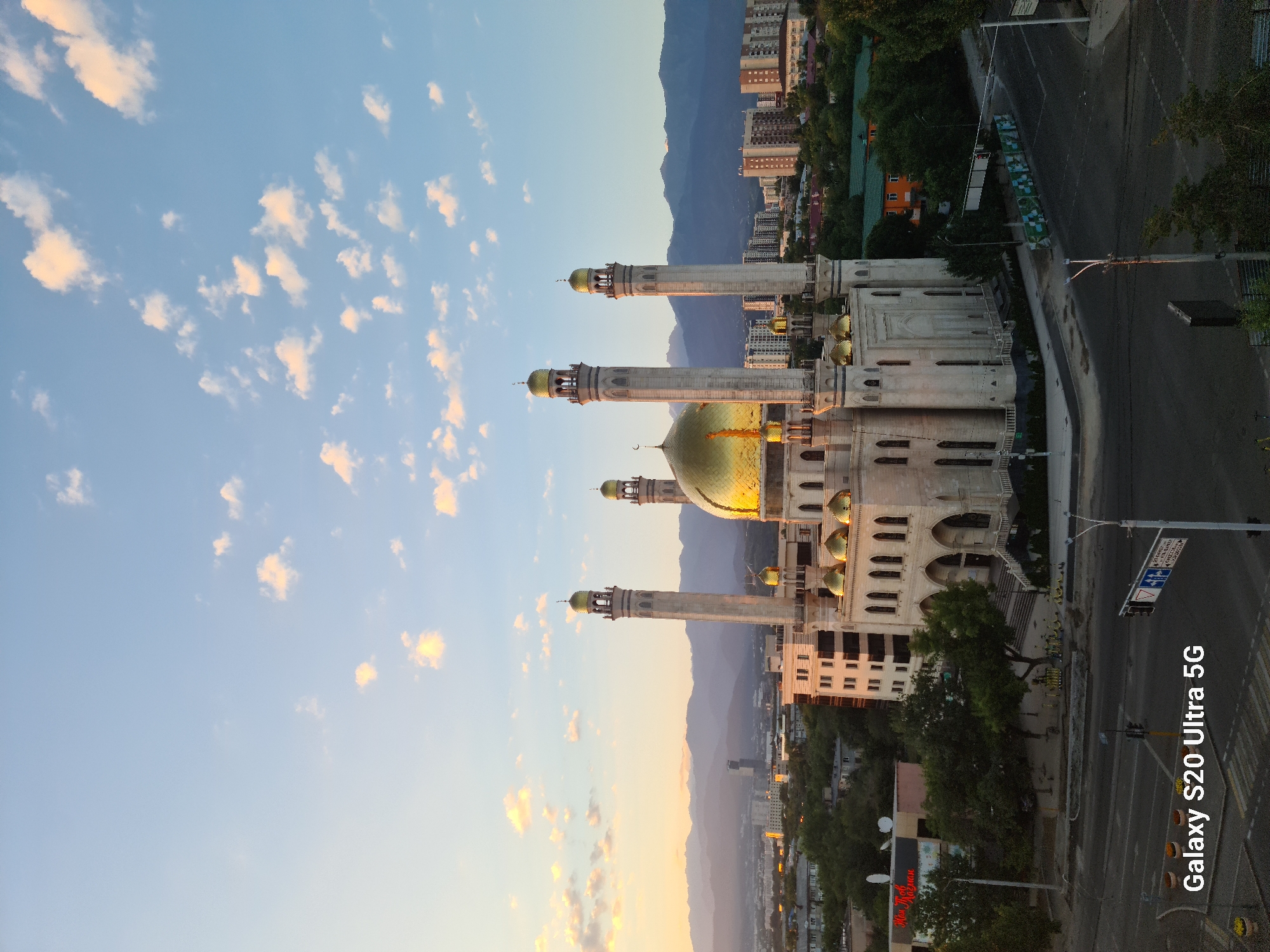 The image captures a panoramic view of the city of Almaty, Kazakhstan, with a prominent mosque as the central subject. The mosque features multiple minarets and a large golden dome, characteristic of Islamic architecture. The building is illuminated by the warm glow of what appears to be either sunrise or sunset, casting long shadows and highlighting the golden domes.
Surrounding the mosque is an urban landscape, including residential buildings, commercial establishments, and dense green trees. In the background, a range of majestic mountains forms a dramatic backdrop against the sky, suggesting the city's proximity to the Tien Shan mountain range. The sky is a gradient of soft blue and orange hues, with scattered clouds, further enhancing the atmospheric quality of the scene.
The foreground shows a street intersection with traffic lights and road markings, indicating a busy urban environment. A sign in the lower left corner reads "Galaxy S20 Ultra 5G," identifying the device used to capture the photograph. No people are clearly visible in the image, suggesting a moment of quiet in the city. The overall impression is one of serene beauty, blending architectural grandeur with natural landscapes.