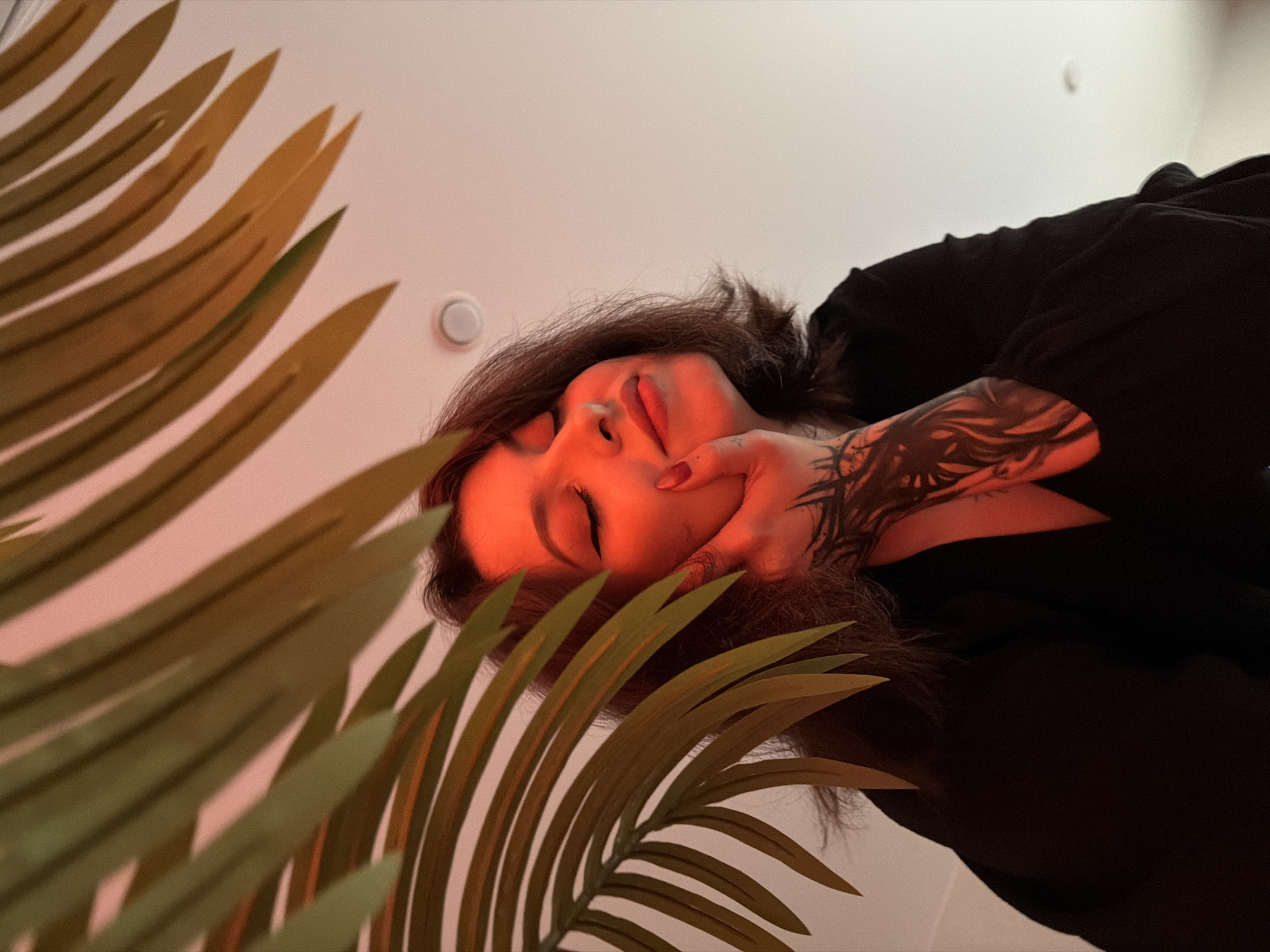 A low-angle medium shot captures a person with their eyes closed, head tilted back slightly, and right hand touching their cheek, framed by green palm fronds under red lighting. The person has short, dark hair and is wearing black clothing. A tattoo is visible on their left forearm. The background is a white ceiling with a circular light fixture. The scene is indoors in an unknown city.