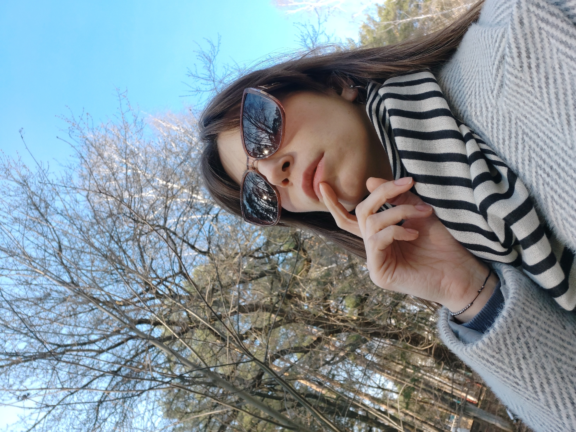 A young woman wearing sunglasses, a gray and white coat, and a black and white striped scarf is posing for a selfie. She is outside, with trees and a bright blue sky in the background. The location is in Chaikovsky, Russia, as can be discerned from metadata. The woman's face is partially obscured by sunglasses, but her mouth is visible as is a hand with a finger raised towards the lips. The reflection in the sunglasses also suggests trees.
