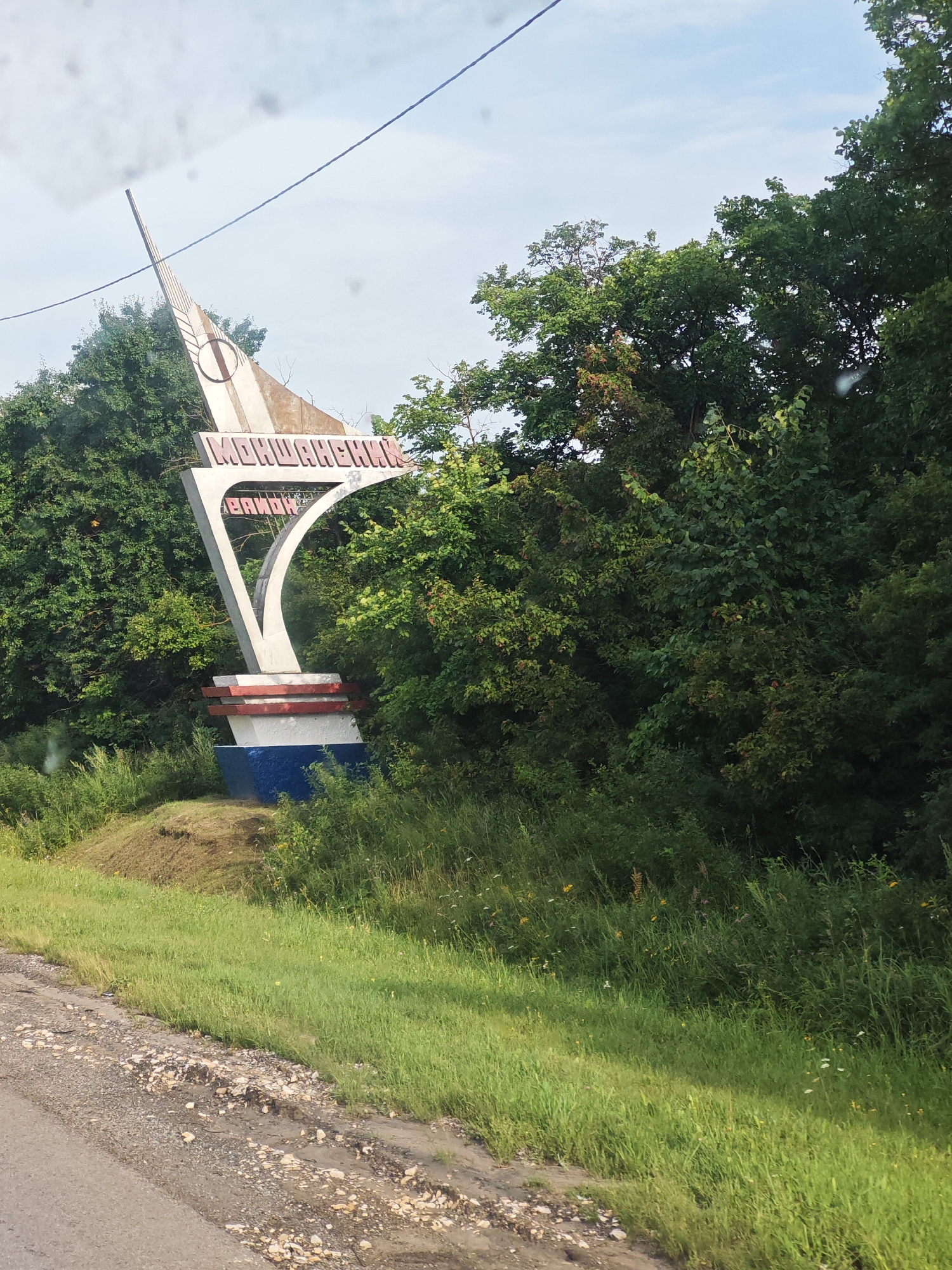 The image shows a landmark sign at the entrance to Shirokois, Russia. The sign is made of concrete and is decorated with red and blue stripes at its base. The top portion of the sign features a stylized depiction of a wheat stalk and a circular emblem. Prominently displayed on the sign in red Cyrillic letters are the words "ШИРОКОИС" (Shirokois) and below it, "РАЙОН" (Rayon), indicating it marks the district. The sign is situated amidst lush green trees and tall grass, suggesting a rural or suburban setting. The road in the foreground is unpaved and rocky. The sky is overcast with a single power line visible.