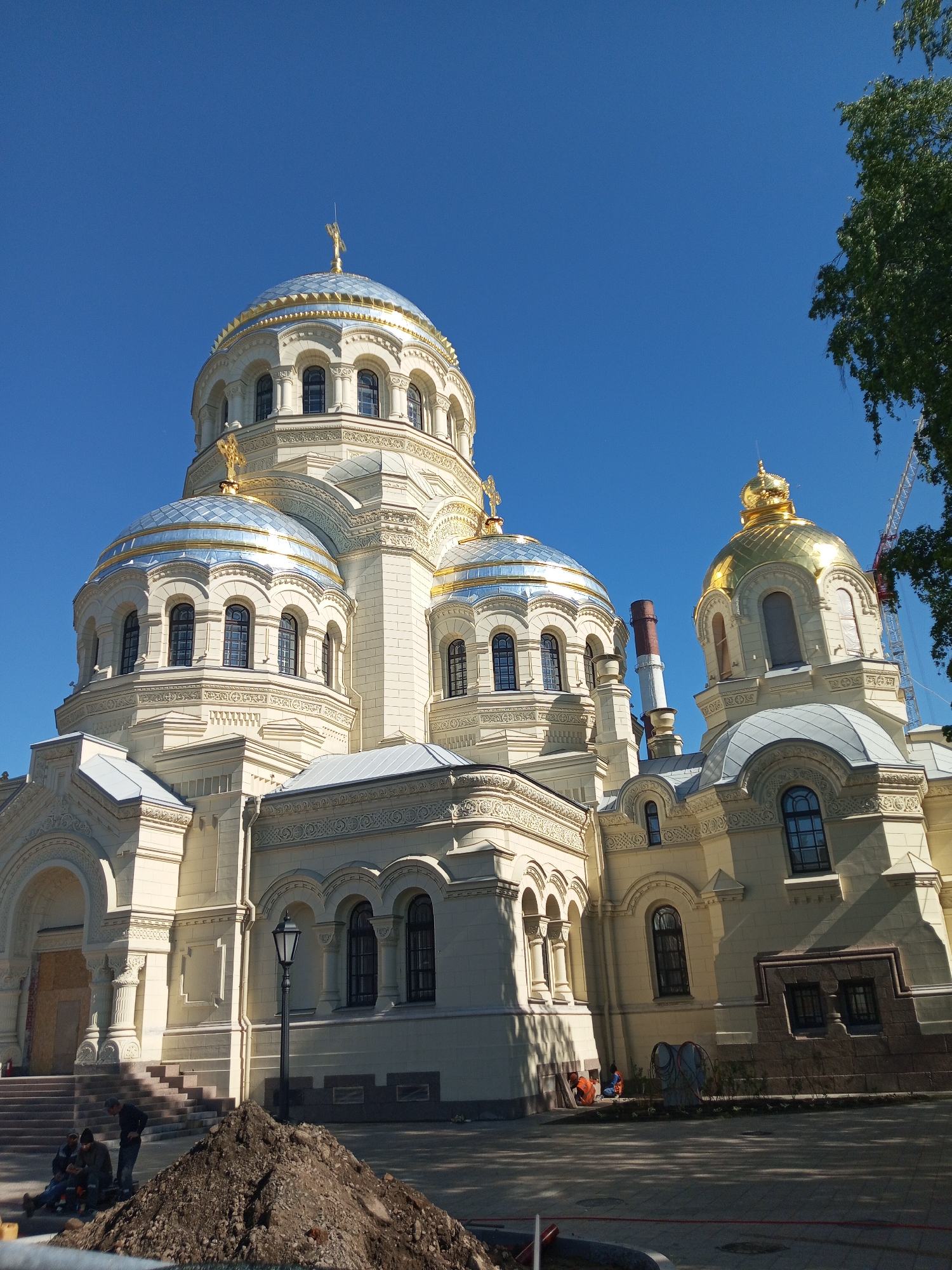 The image shows a large, ornate cathedral in the city of Saint Petersburg, Russia, under a clear blue sky. The cathedral is primarily white with multiple domes, some covered in silver and others in gold. Each dome is topped with a golden Orthodox cross. There are numerous arched windows and decorative architectural details.
In the foreground, there is a pile of dark soil and a group of three people, one standing and two sitting, possibly resting or working. Further towards the building, near the base, are two people in what appears to be work clothes.
To the right of the cathedral, a construction crane is visible. A lamppost stands near the front of the cathedral.