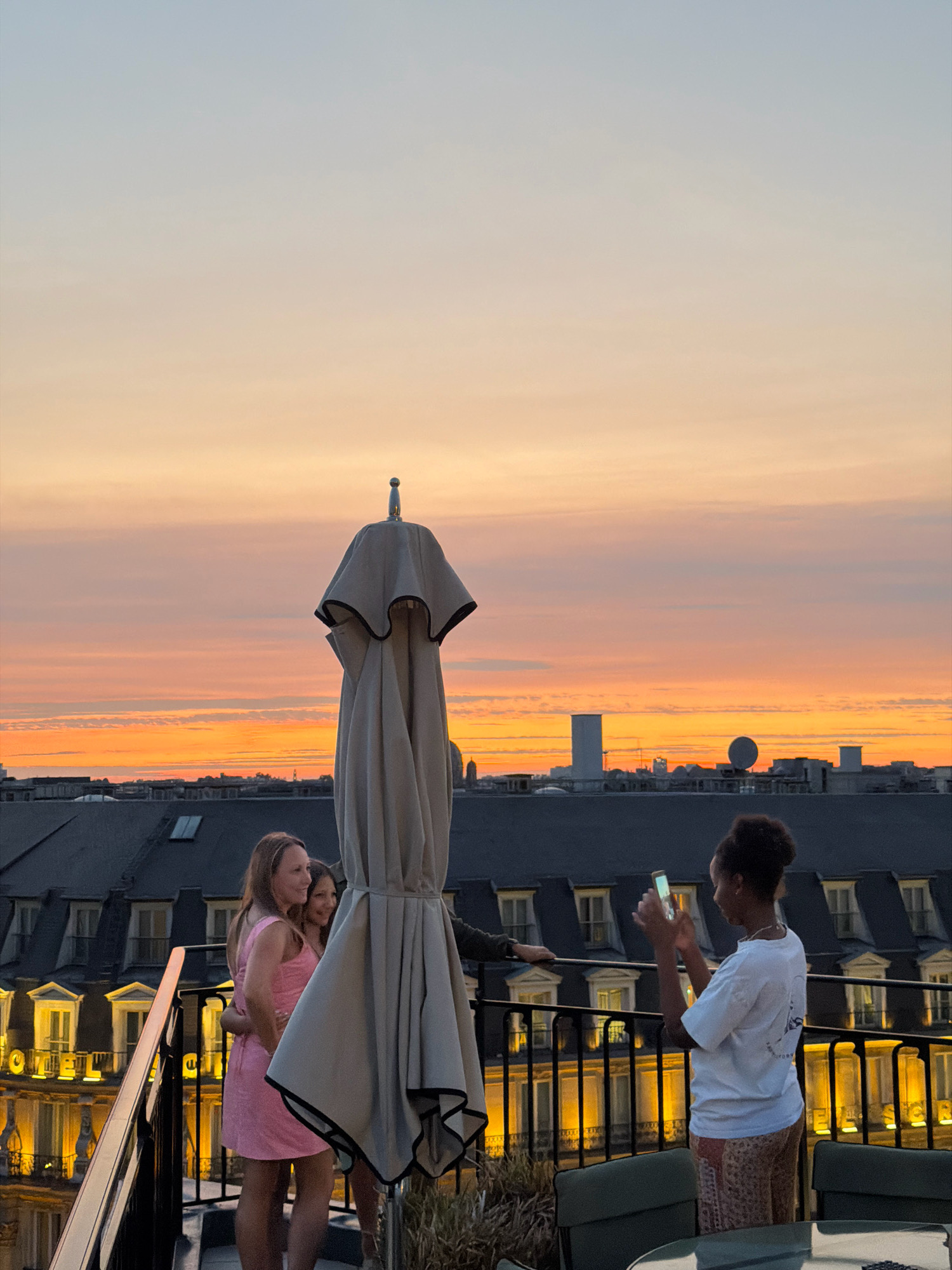 This image depicts a rooftop scene in Paris, France, during a sunset. In the foreground, three young women are present. Two of them, positioned on the left, are embracing and smiling. The woman on the left wears a pink dress, and the woman next to her, partially obscured, is also in a pink garment. A large, folded beige umbrella stands prominently in the center of the frame. On the right, a third woman, wearing a white t-shirt and patterned pants, is taking a photograph with her phone. In the background, the Parisian cityscape is visible, characterized by dark, ornate rooftops and illuminated windows of buildings. The sky displays a gradient of warm sunset colors, transitioning from orange and pink near the horizon to a soft blue overhead. The overall atmosphere suggests a relaxed and scenic evening.