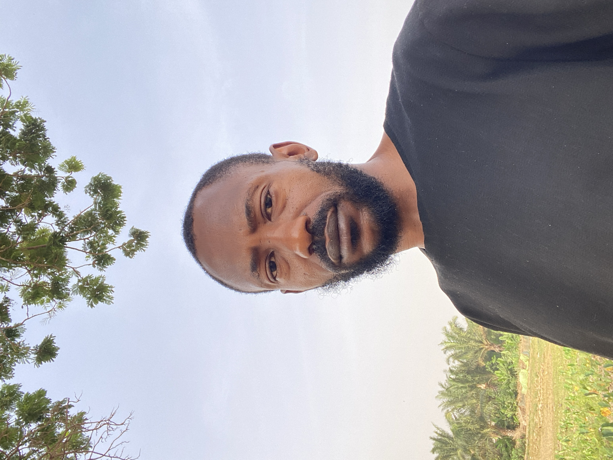 A man with dark skin, a full beard, and a shaved head is looking directly at the camera. He is wearing a black shirt. The sky is overcast. In the background, there is a tree with green foliage on the left side of the frame and palm trees and vegetation on the right. The photograph appears to have been taken from a low angle. There is no indication of being in Sabon Tasha, Nigeria.