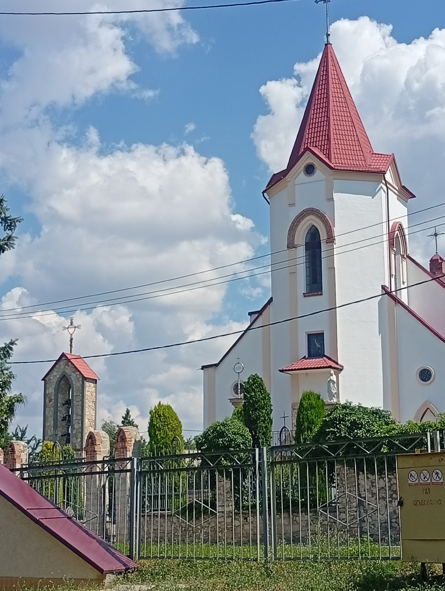 The image shows a bright, sunny day in Rybnitsa, Moldova. The foreground features a metal fence and a low, angled roof of a building. Behind the fence, there is a garden with green grass and various shrubs and trees. In the mid-ground, a stone structure with a red roof, likely a bell tower, stands to the left, topped with a cross. To the right of the bell tower, a white church with a prominent red-tiled spire dominates the scene. The church has arched windows and a statue of a figure, possibly a religious icon, is visible on a small pediment. Power lines are strung across the sky, which is partly cloudy with large white cumulus clouds against a blue sky. A yellow electrical box is attached to the fence on the right side of the image, displaying warning symbols and text. The overall impression is one of a peaceful, somewhat rural religious site on a clear day.