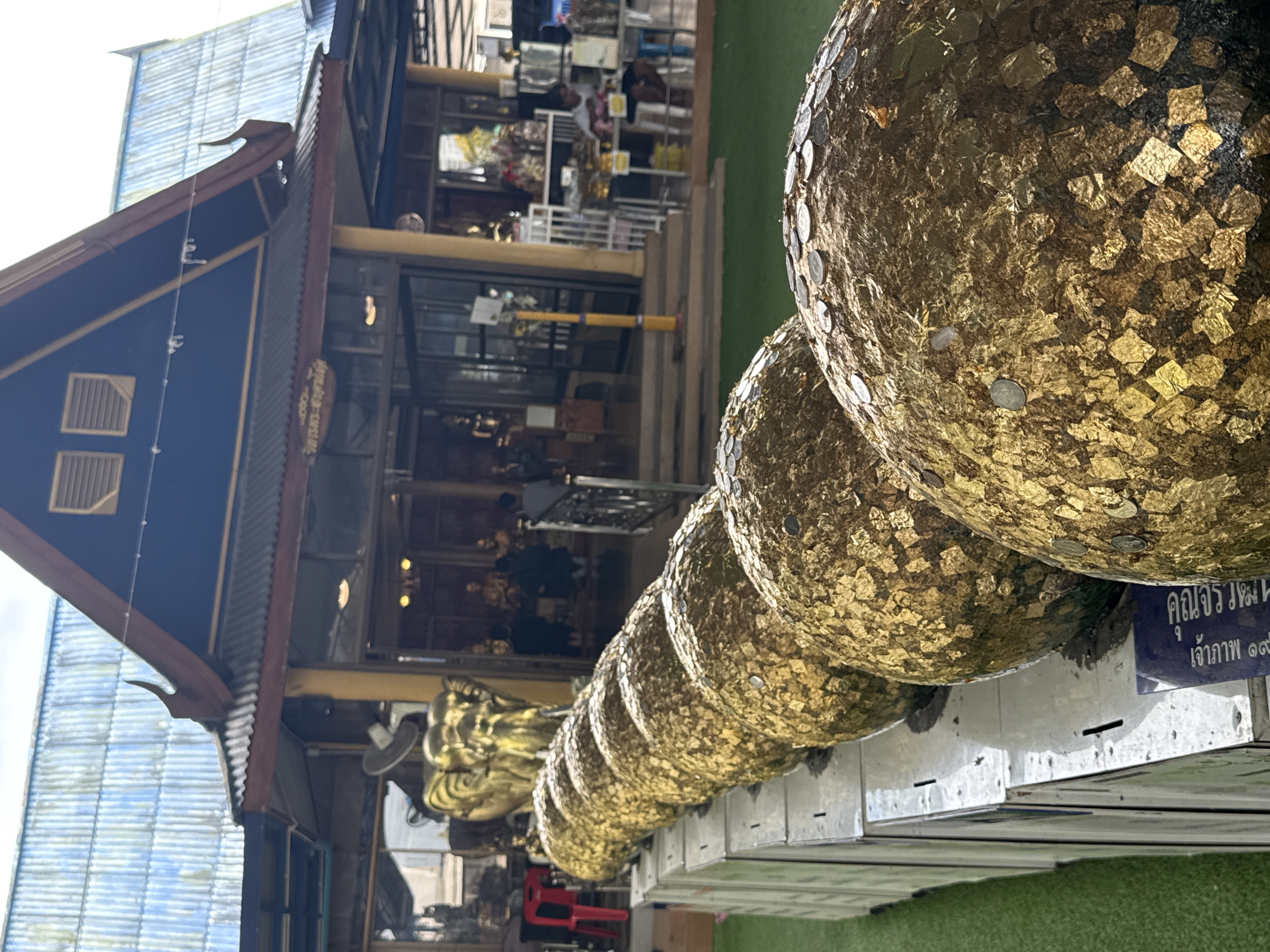 A row of large, spherical objects covered in gold leaf and coins are lined up in the foreground of the image. These objects are placed on a white metal structure, with a green carpeted area to the left. In the background, a building with a dark blue roof and yellow pillars is visible. The building appears to be a temple or shrine, with statues and offerings inside. Several people are seen inside and around the building, some of whom appear to be visitors. The sky is overcast, suggesting a cloudy day. The image appears to have been taken in Tambon Na Chom Thian, Thailand, given the architectural style and the context provided.