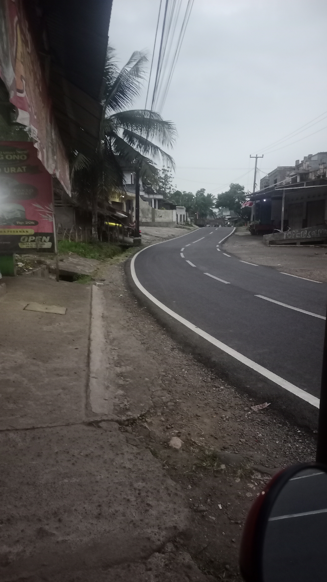 The image depicts a street scene in Cogreg, Indonesia, under an overcast sky, possibly during dawn or dusk. A paved road curves into the distance, with white dashed lines marking the lanes. The road is bordered by a gravel shoulder and a concrete sidewalk on the left.
On the left side of the road, a building with a dark awning is visible, featuring a large banner with red and white text. Partially visible text on the banner reads "GONO URAT" and "OPEN". Adjacent to this building is a palm tree with its fronds extending over the scene.
On the right side of the road, several buildings are visible, some with pitched roofs. One building has an awning with lights illuminating it. The architecture suggests a mix of residential and commercial structures. A utility pole with wires is also present. The ground is a mixture of dirt, gravel, and scattered debris. In the foreground, a portion of a reflective surface, possibly a mirror of a vehicle, is visible at the bottom right of the image. No people are clearly visible in the scene. The overall impression is of a quiet, somewhat rural street on the outskirts of a town.