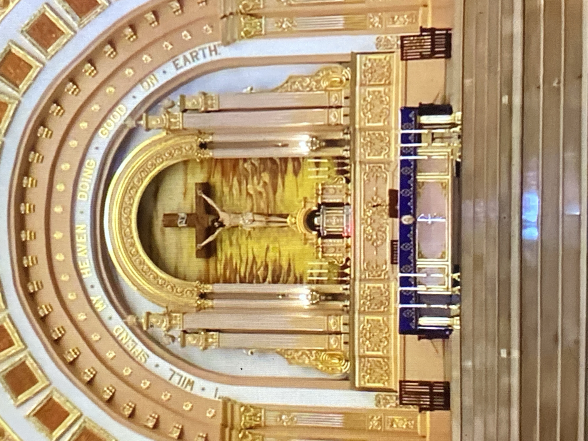 This indoor shot focuses on the altar inside the church in Tinaquillo, Venezuela. The altar is decorated with an image of the crucifixion in the middle. Above it, text is inscribed, stating, "My heaven doing good on earth I will spend." The altar is also decorated with gold and white colors, creating a rich and spiritual ambiance. To the right of the altar, a plain, light-colored wall adds contrast to the decorated space. The shot is taken from above, offering a clear view of the layout of the church.