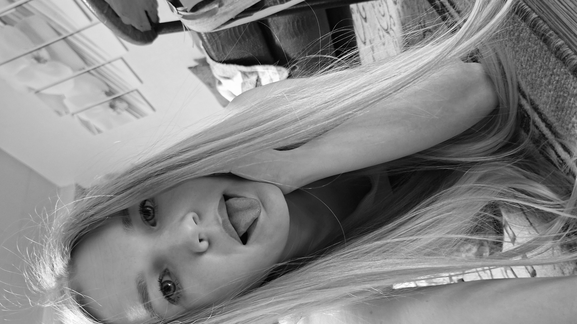 A black and white close-up photograph shows a young woman with long, straight, light-colored hair sticking her tongue out. Her eyes are visible, and she appears to be indoors. The background is blurry, but there are hints of furniture and a window. The setting may be a residential space, but no definitive information about the location can be determined from the image itself. The photograph does not provide any information that verifies the location is in Большой Букор, Russia.