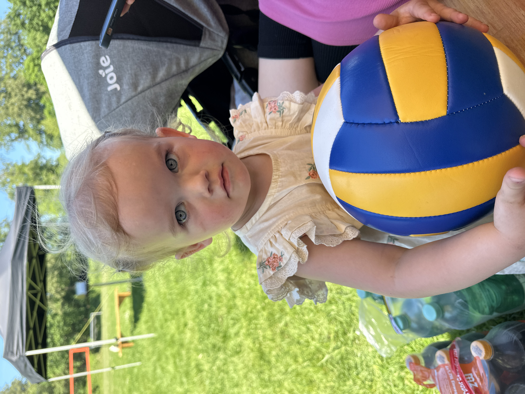 A young child with blonde hair and blue eyes sits in a grey stroller on a grassy area in Grünberg, Germany. The child wears a light-colored dress with floral embroidery. In her hands, she holds a blue, yellow, and white ball. Bottles of water and other beverages are on the grass beside her. A black tent-like structure is visible in the background.