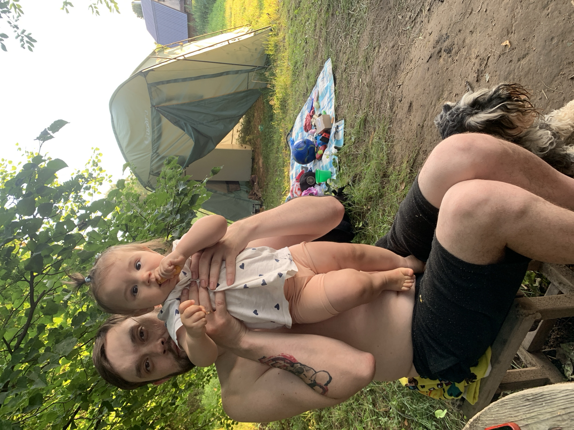 In Zasechnyi, Russia, a bare-chested man with a beard and tattoos sits on a wooden stool, holding a baby. The baby, wearing a white shirt with blue hearts and peach-colored shorts, is eating. Behind them, a green tent is set up on a grassy area with a checkered blanket, toys, and a ball scattered on it. A small, shaggy dog lies on the ground to the man's right. The scene is outdoors, with trees and a house visible in the background, suggesting a camping or picnic setting.