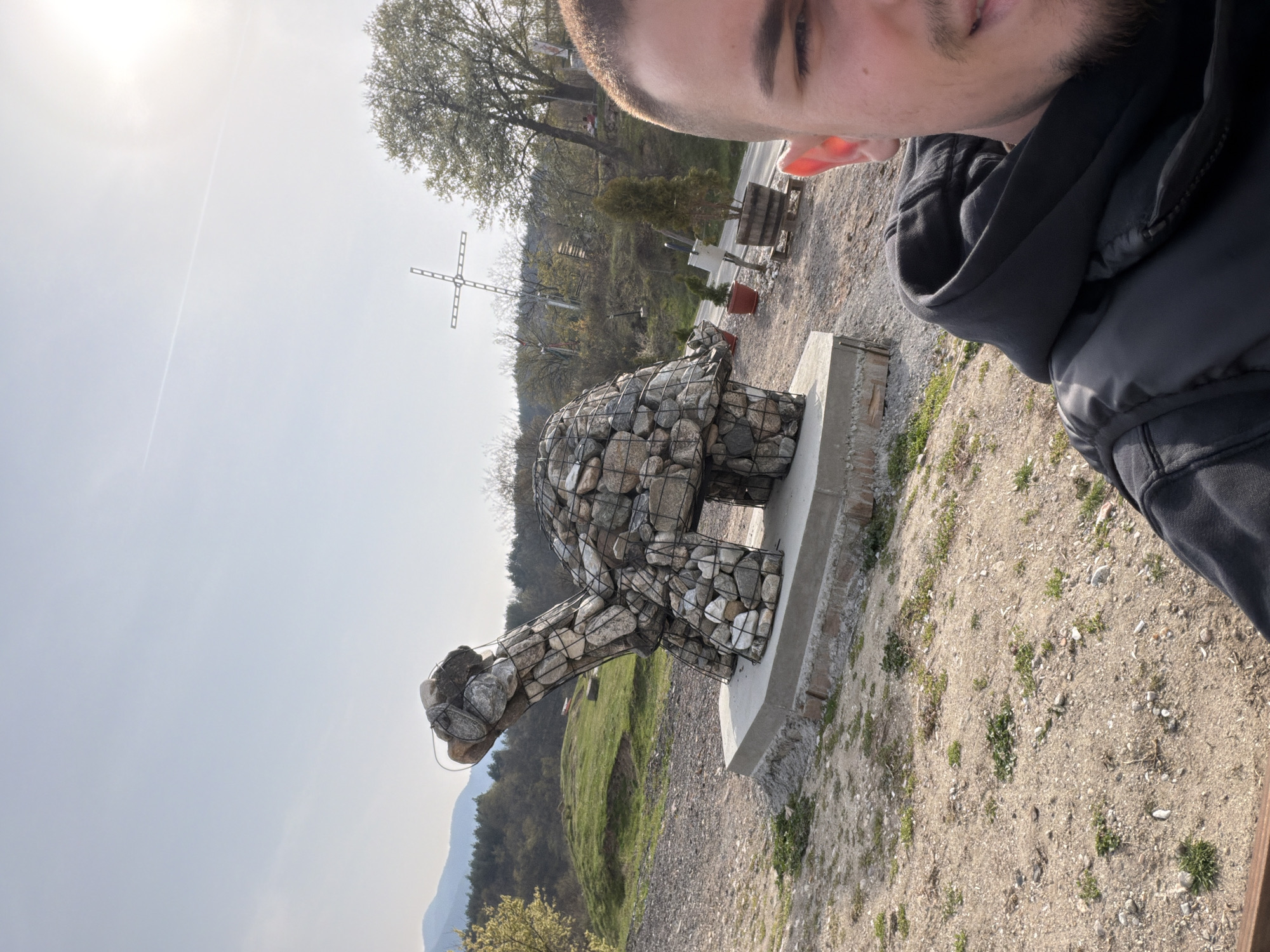 A selfie captured in the area of Sveti Konstantin, Bulgaria, shows a man with short hair and a beard wearing a dark hooded jacket, positioned in the upper right of the frame. A large, artistic stone turtle sculpture stands on a concrete base in the center of the image. In the background, a metal cross is visible on a hill, surrounded by trees and vegetation. The environment consists of a gravel area with sparse plant growth. Mountains are visible in the distance under a bright sky.