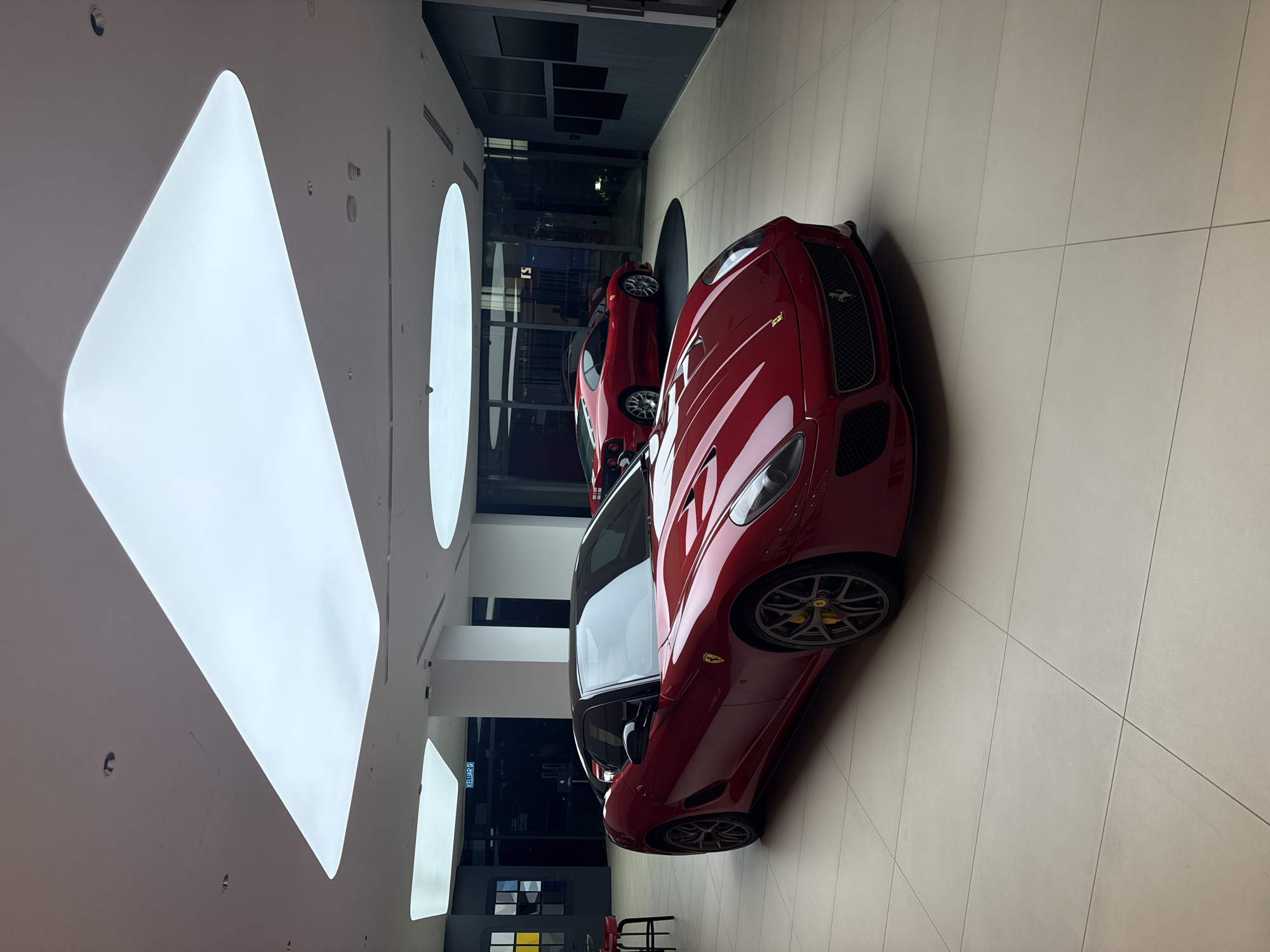 The image captures a car dealership setting in Kuala Lumpur, Malaysia. Two red Ferrari sports cars are prominently displayed on a light-colored tiled floor, positioned in a way that allows the first to be partially visible in the background. The cars are positioned beneath artificial ceiling lights and are near transparent storefront windows. Minimal furnishings such as chairs are present in the space. Signage in the storefront window identifies the location of the dealership.
