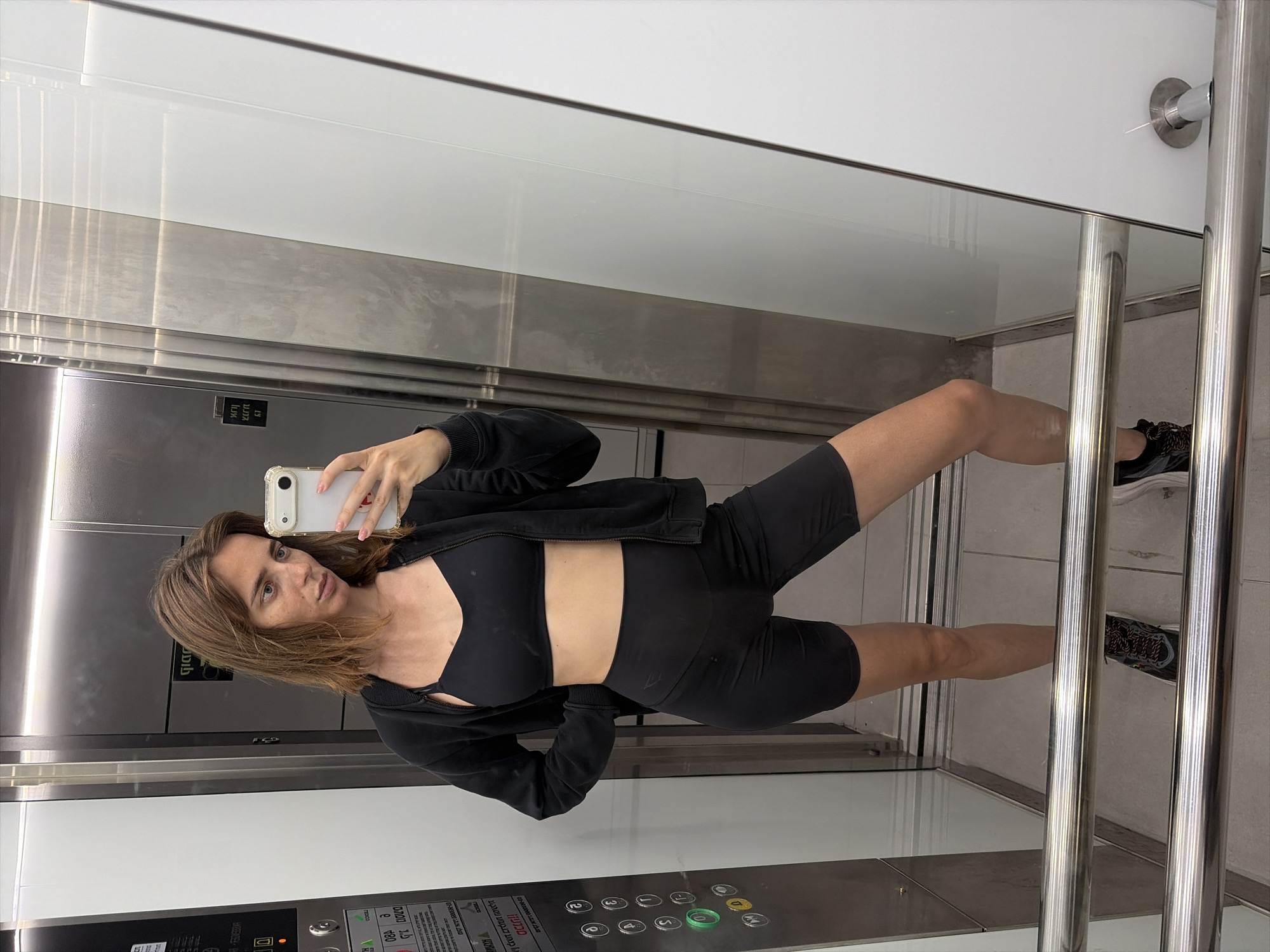 A woman stands in an elevator, taking a mirror selfie. She is wearing a black sports bra, black biker shorts, and a black zip-up hoodie. Her hair is medium brown and styled in loose waves. She is holding a white smartphone in her right hand. The elevator has stainless steel walls and a tiled floor. Buttons are visible on the left wall of the elevator. The setting is an elevator in Tel-Aviv, Israel.