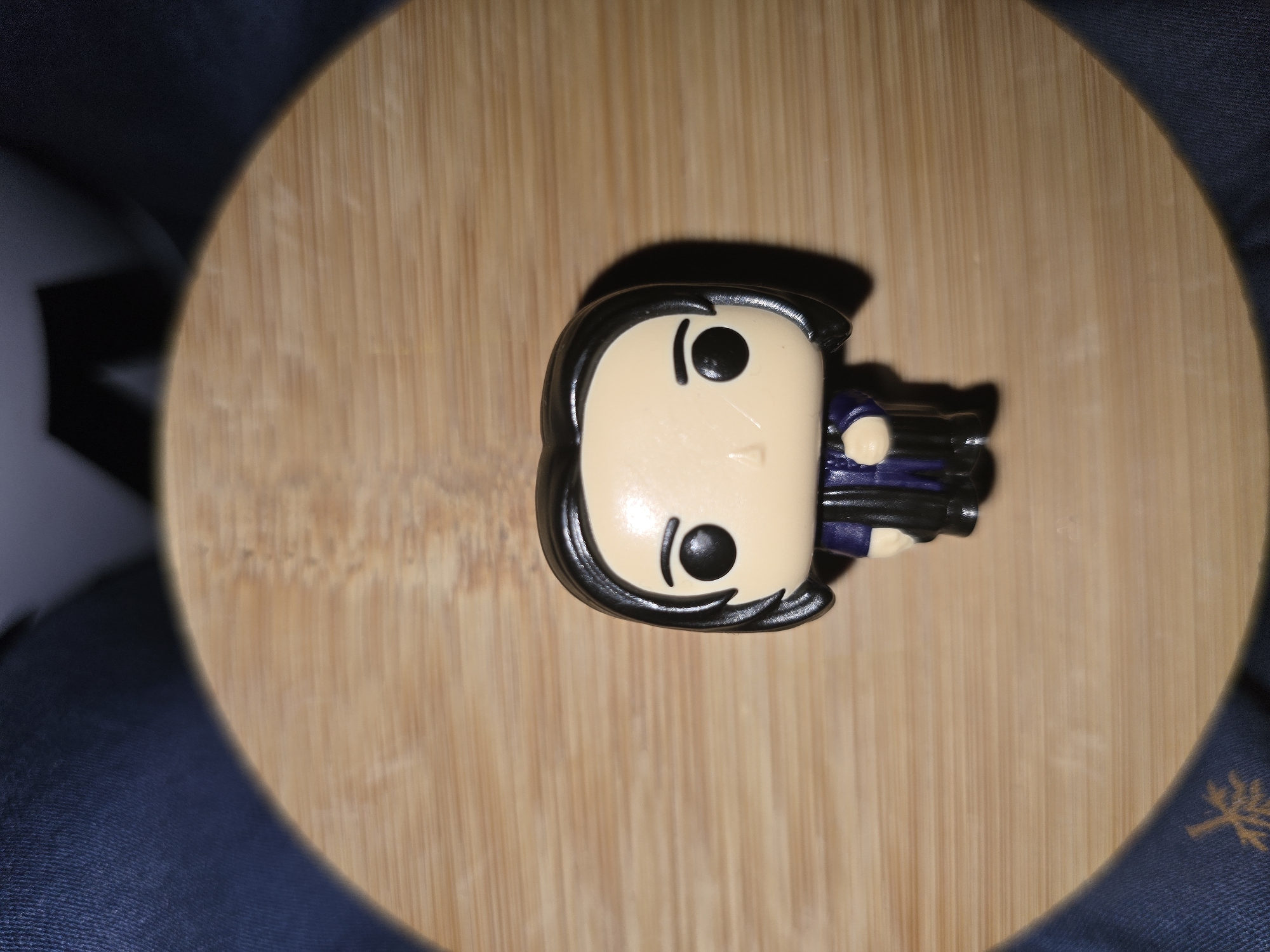 A Funko Pop figure of Severus Snape from Harry Potter stands on a light brown wooden surface. The figure is dressed in a dark purple robe with black trim. The figure has black hair, pale skin, black eyes, and a neutral expression. The background consists of dark blue fabric. The scene is not located in Petropavlivska Borshchahivka, Ukraine.