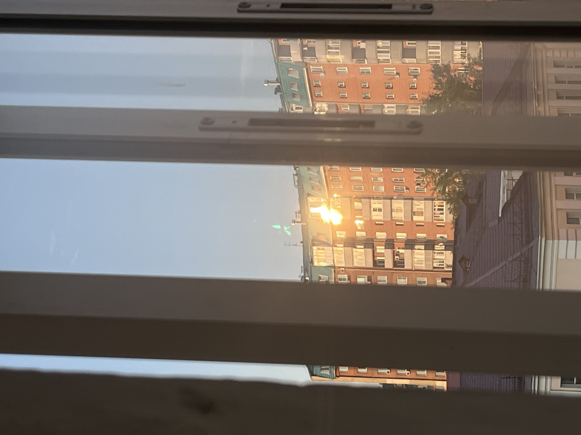The image shows a view of apartment buildings and rooftops through a window frame. The sky is clear and blue, suggesting it is daytime. The sun is setting or rising, casting a strong golden light on the buildings, creating a bright flare in the center of the image. Some of the apartment buildings are orange brick, while others appear to be gray or white. The rooftops are dark and sloped. There are trees visible between some of the buildings. The window frame has vertical white mullions that divide the view. There are no people visible in the image.
