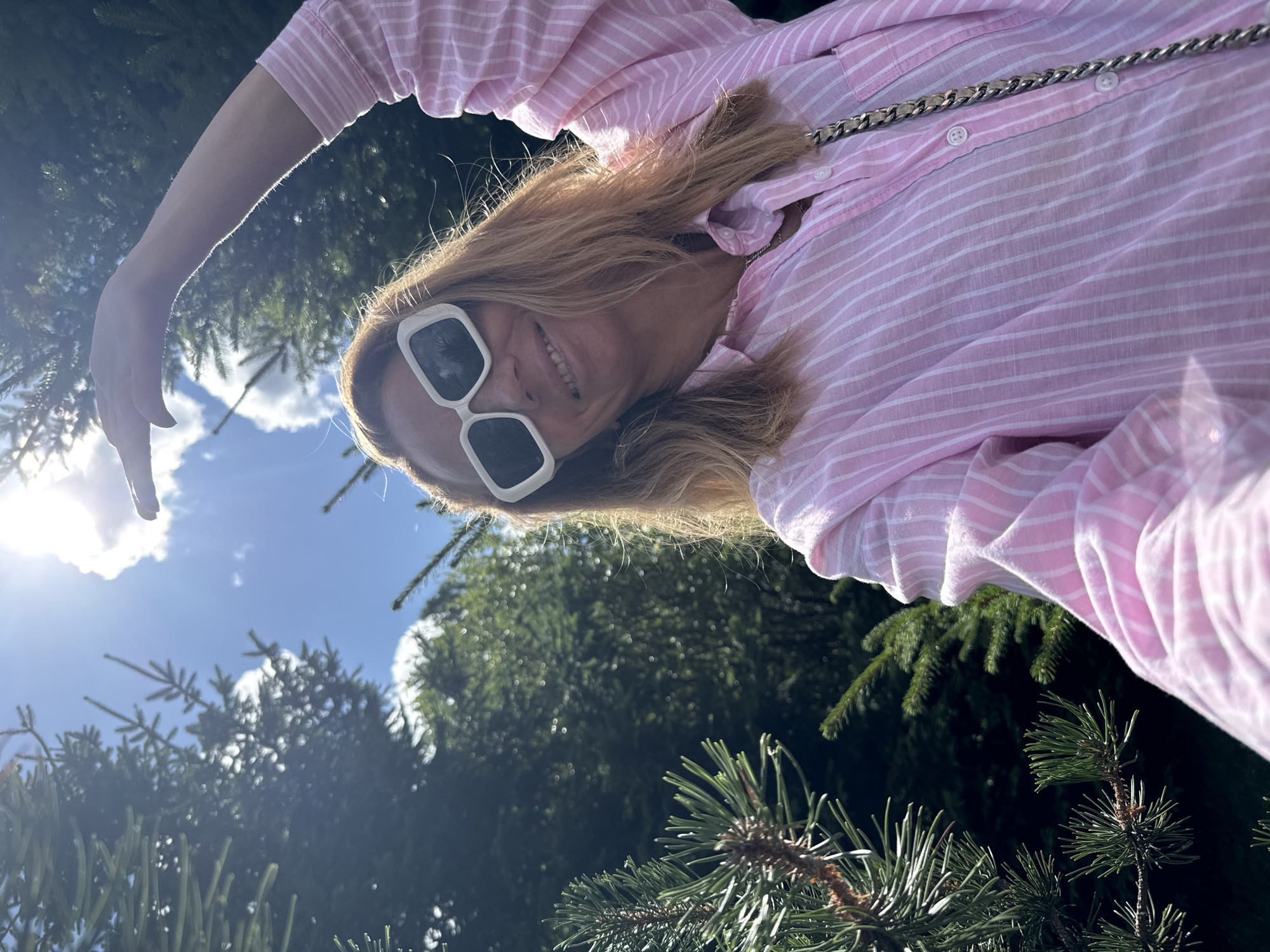 A medium shot photograph, taken outdoors during the daytime, shows a woman with shoulder-length blonde hair, wearing large white rectangular sunglasses and a pink and white striped button-up shirt. She is smiling and raising her right arm towards the sky. Behind her, numerous evergreen trees are visible, with a bright sky and sun shining through the branches. The location is identified as being in the city of Sinyavino, Russia.