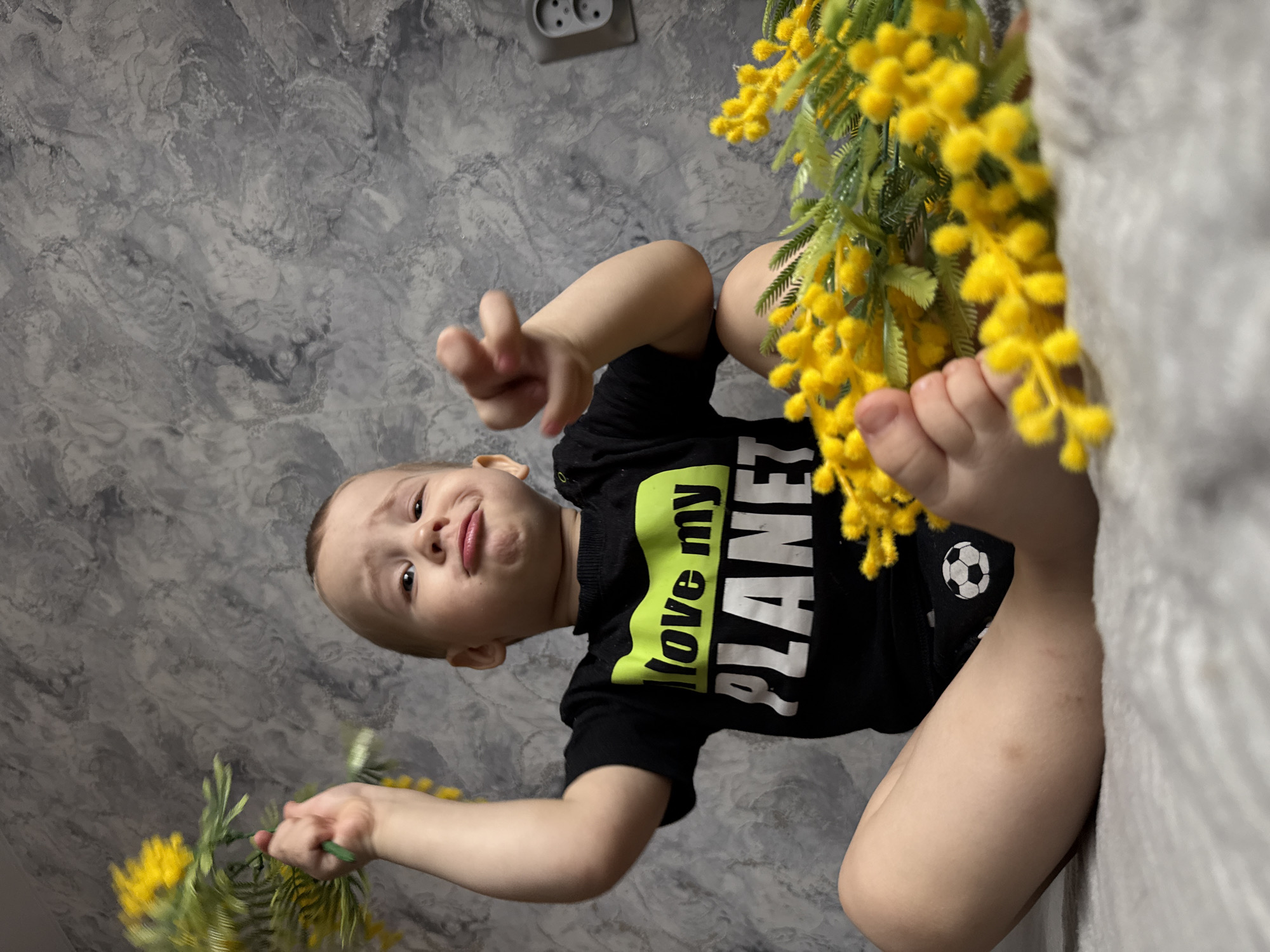 A young child is sitting on a bed in a room with grey marble-patterned walls, possibly in Rusanovo, Russia. The child is wearing a black shirt with the phrase "Love my PLANET" in green and white lettering and a soccer ball design. The child is holding a bouquet of yellow flowers in one hand, and another bouquet is near their feet. An electrical outlet is visible on the wall.