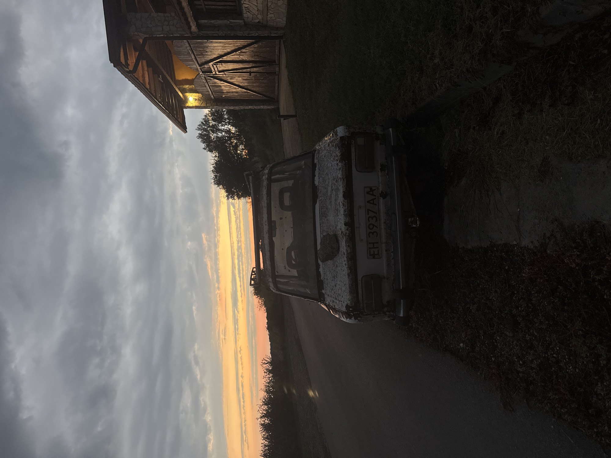 The image depicts a rural roadside scene in Radishevo, Bulgaria, at dusk. An old, weathered car, possibly a Lada, is parked on the shoulder of a paved road. The car is covered in dirt and appears to be in disrepair. Its license plate, partially obscured, reads "EH 3937 AA". In the background, a rustic building with a large wooden gate and an illuminated lamp stands to the right. The sky is filled with dramatic clouds tinged with the orange and yellow hues of a sunset. Trees and low-lying vegetation line the road and surround the property. The overall atmosphere is one of stillness and abandonment.
