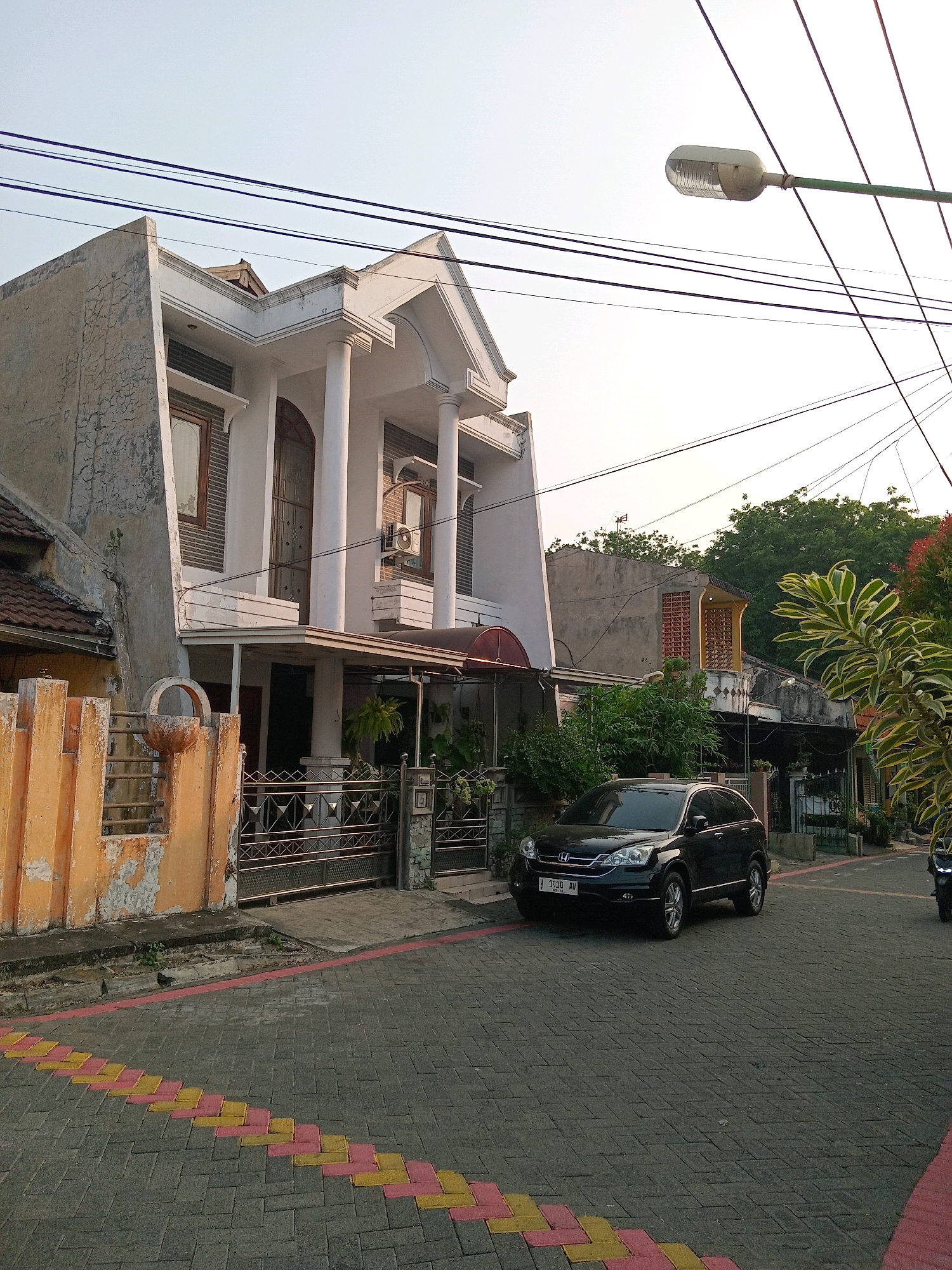 The image depicts a street scene in the city of Randuagung, Indonesia, with a focus on residential architecture and a parked car. In the foreground, a paved street with a decorative pattern of yellow and pink bricks runs diagonally. A black SUV is parked on the right side of the street. The background features a two-story house with a white facade, classical columns, and arched windows. An air conditioning unit is visible on the exterior wall of the house. To the left of the house, a fence with orange-painted posts and metal bars separates the property from the street. Overhead, power lines crisscross against a pale sky, and a street lamp is mounted on a pole. Lush green foliage, including a large plant with variegated leaves, adds a natural element to the urban environment. The overall impression is of a quiet residential street during daylight hours.