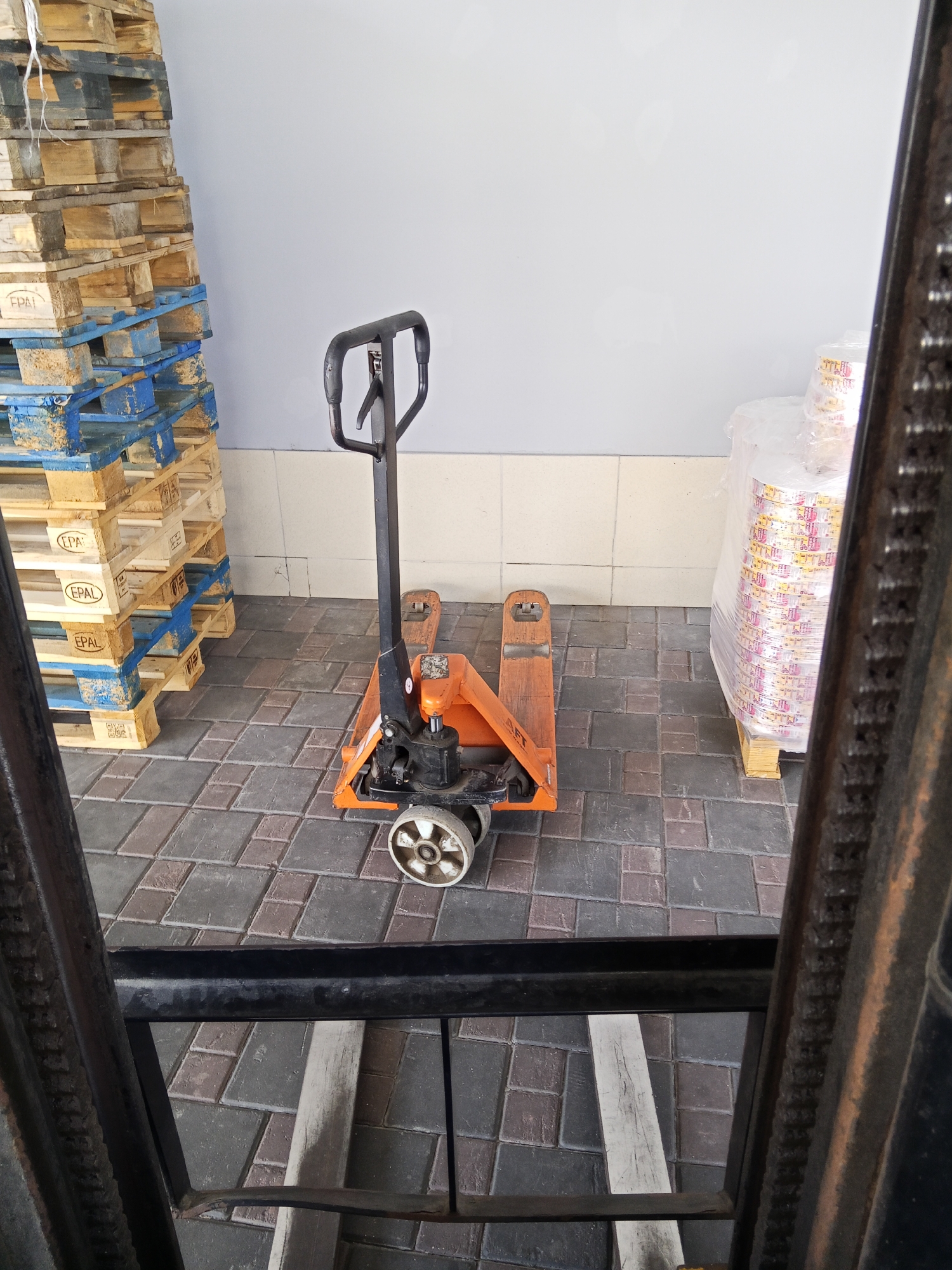 An orange pallet jack with a black handle sits on a patterned tile floor in front of a stack of wooden pallets. The pallets are stacked high and are bound with blue plastic straps. To the right of the pallet jack, a large, clear plastic-wrapped bundle of items sits on a wooden pallet. The background is a plain, light-colored wall. The foreground shows the metal structure of what appears to be a forklift. The scene is indoors, possibly in a warehouse or storage area. The city of Ceadîr-Lunga, Moldova, is not depicted in the image, and there is no information within the image to suggest its location.