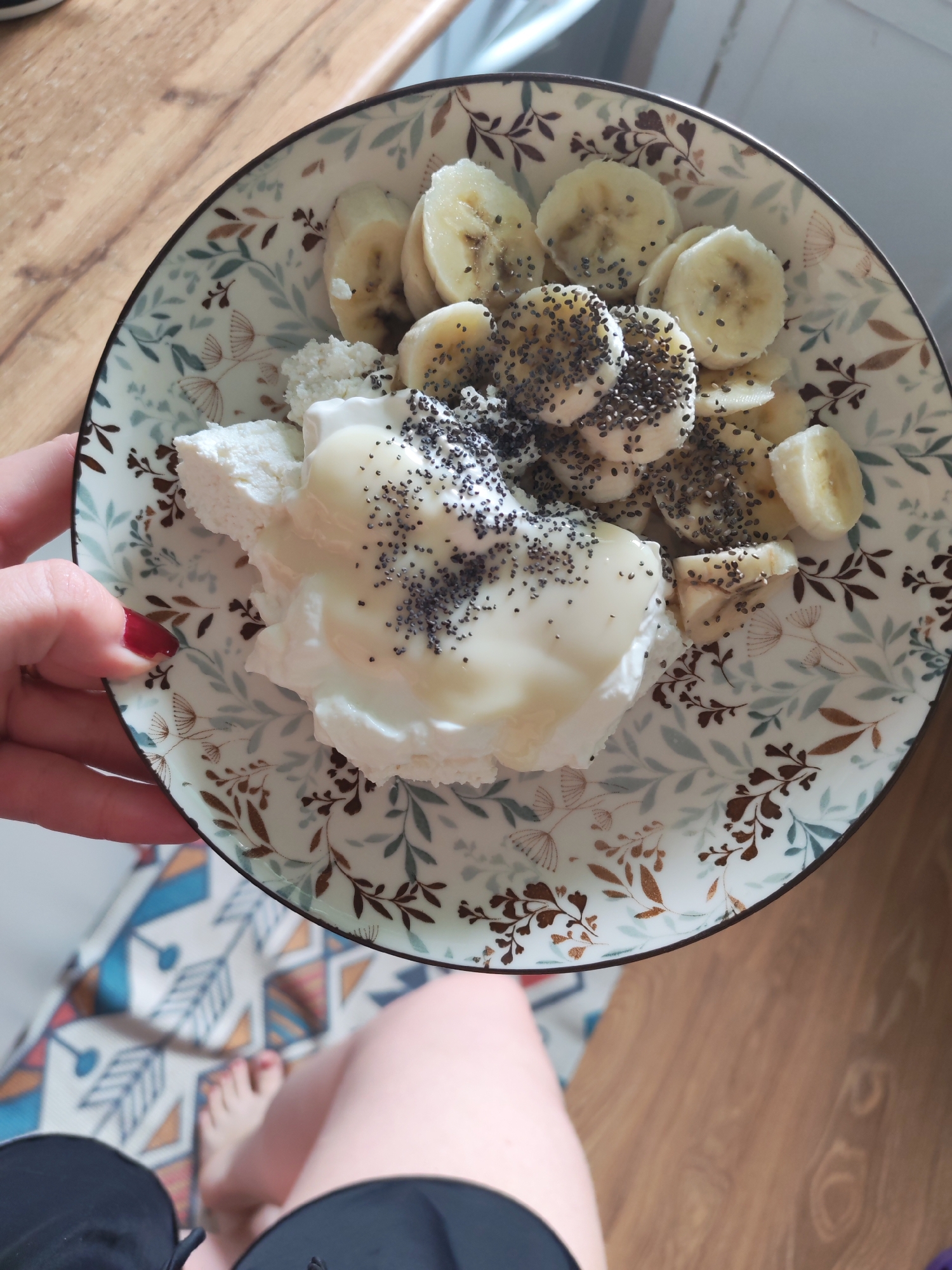 A close-up, overhead shot shows a person's hand holding a patterned bowl filled with a breakfast dish. The dish consists of cottage cheese, sliced bananas topped with chia seeds, and a drizzle of a light-colored sauce, possibly yogurt or honey. The bowl is decorated with a floral pattern in shades of brown, blue, and green. The background is a wooden surface. The bottom part of the image shows a person's legs and feet, as well as a portion of a patterned rug. The scene appears to be indoors, likely in a kitchen or dining area. The location in Izhevsk, Russia, is not discernible from the image itself.