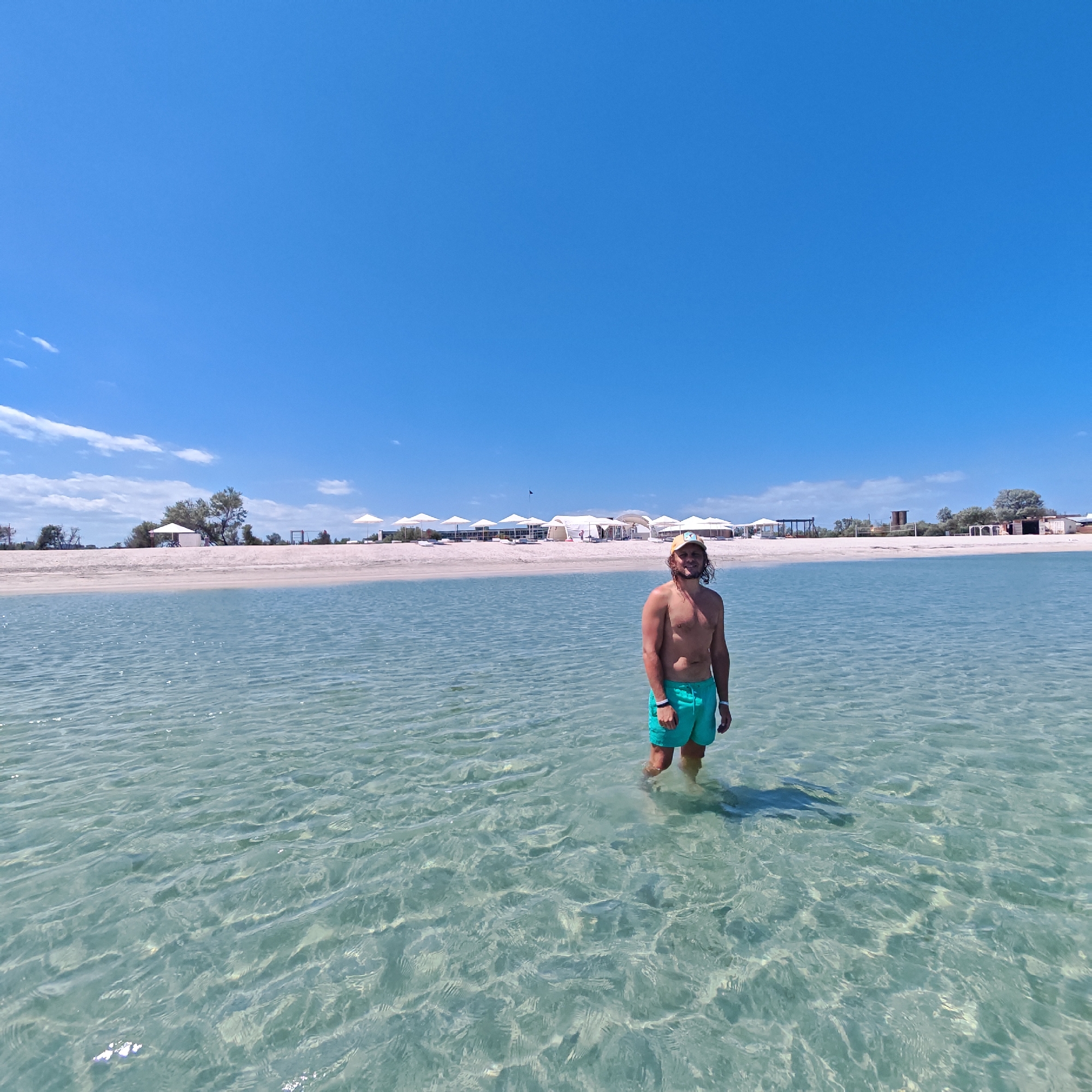 A man stands in clear, shallow water. He is wearing green shorts and a cap. Behind him is a sandy beach with numerous white tent-like structures. The sky is blue with few clouds. The location is likely in городское поселение Щелкино, Ukraine, given the context. The man is standing still in the water. The environment appears to be a beach on a sunny day.