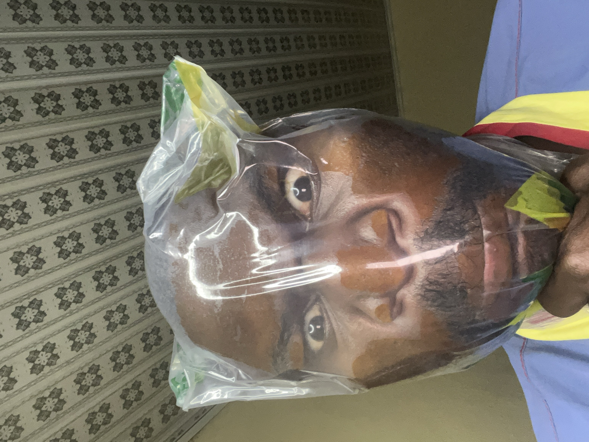 A human head is encased in a clear plastic bag. The individual has dark skin, black hair, and visible facial features. The person wearing a blue shirt with a yellow and red band around the neck. The background consists of a wall with a repeating geometric pattern on the left and a plain, light-colored wall on the right. The photograph was taken in Gura Top, Nigeria.