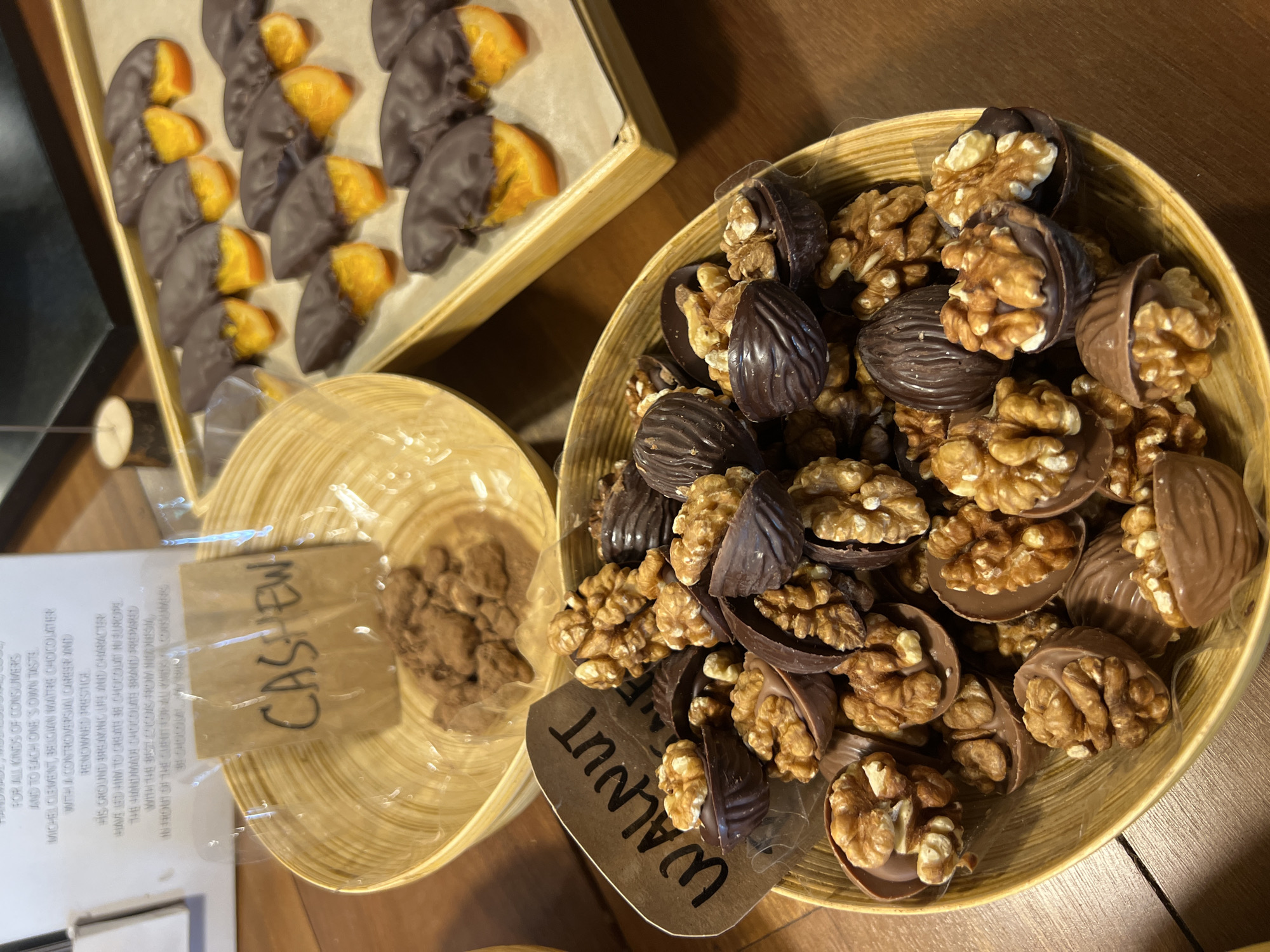 The image shows a close-up of several bowls and a wooden tray filled with chocolate-covered nuts and orange slices. The bowls are made of a light-colored wood and contain what appears to be chocolate-covered walnuts and cashews, with hand-written labels indicating the nut type. The walnuts have their halves dipped in chocolate. The orange slices in the tray have also been dipped in chocolate. The scene is indoors, likely at a store or market selling artisanal chocolates. There are no people visible in the image. The text on the left edge of the image is product information in what is presumably the location of a store selling these products.