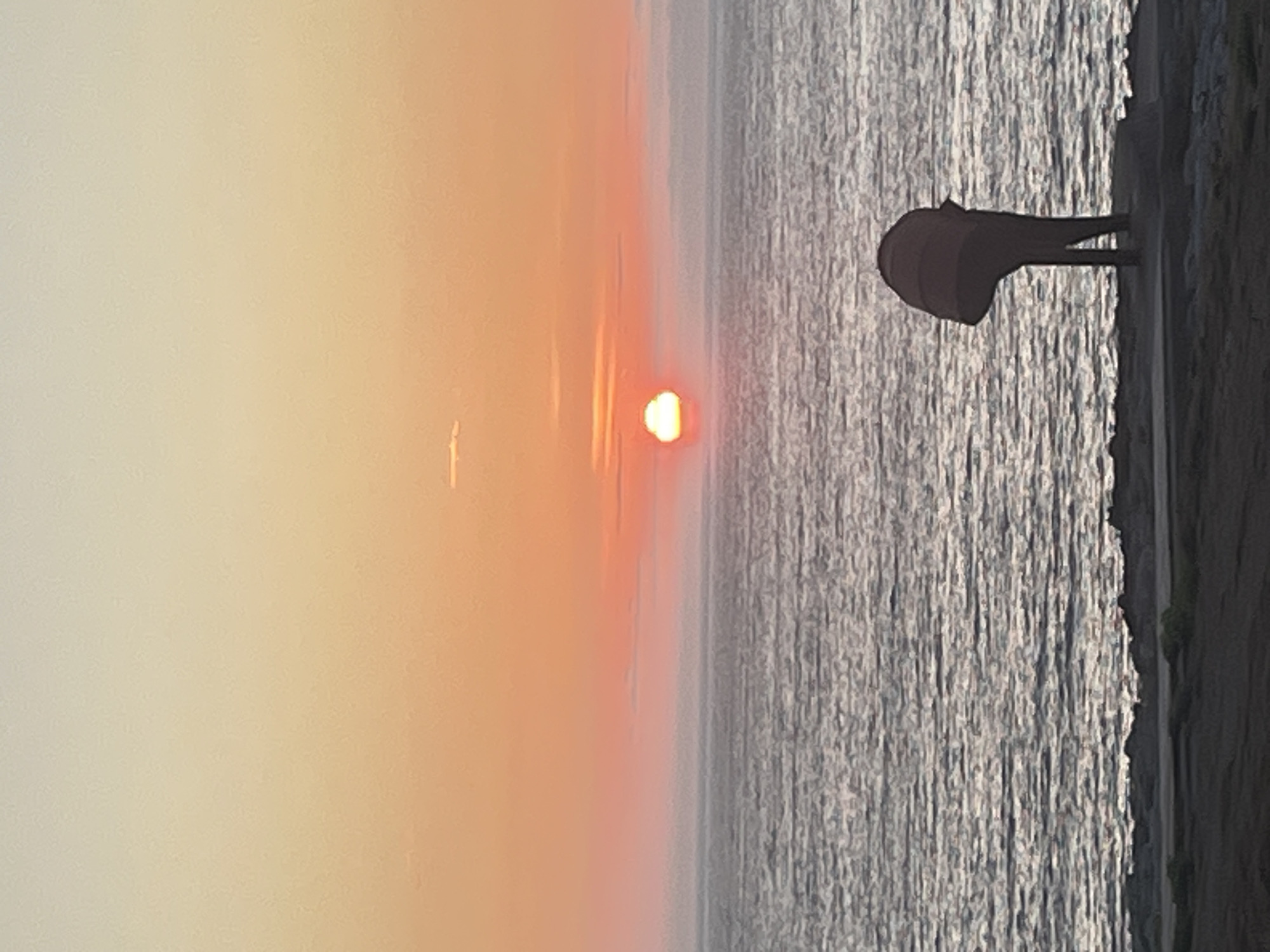 The image depicts a sunset over the Atlantic Ocean, viewed from the coast of A Coruña, Spain. In the foreground, a dark, abstract sculpture with a rounded top and two thin legs stands on a paved area. The sculpture is silhouetted against the sea, which has a textured, rippling surface reflecting the soft colors of the sky. The sky itself transitions from a pale yellow at the top to a vibrant orange and pink near the horizon, where the sun is setting. A thin, horizontal cloud is visible in the upper part of the sky, and a slightly thicker band of clouds runs along the horizon. The coastline in the immediate foreground appears rocky and uneven, with some low-lying vegetation. The overall scene is serene and atmospheric.