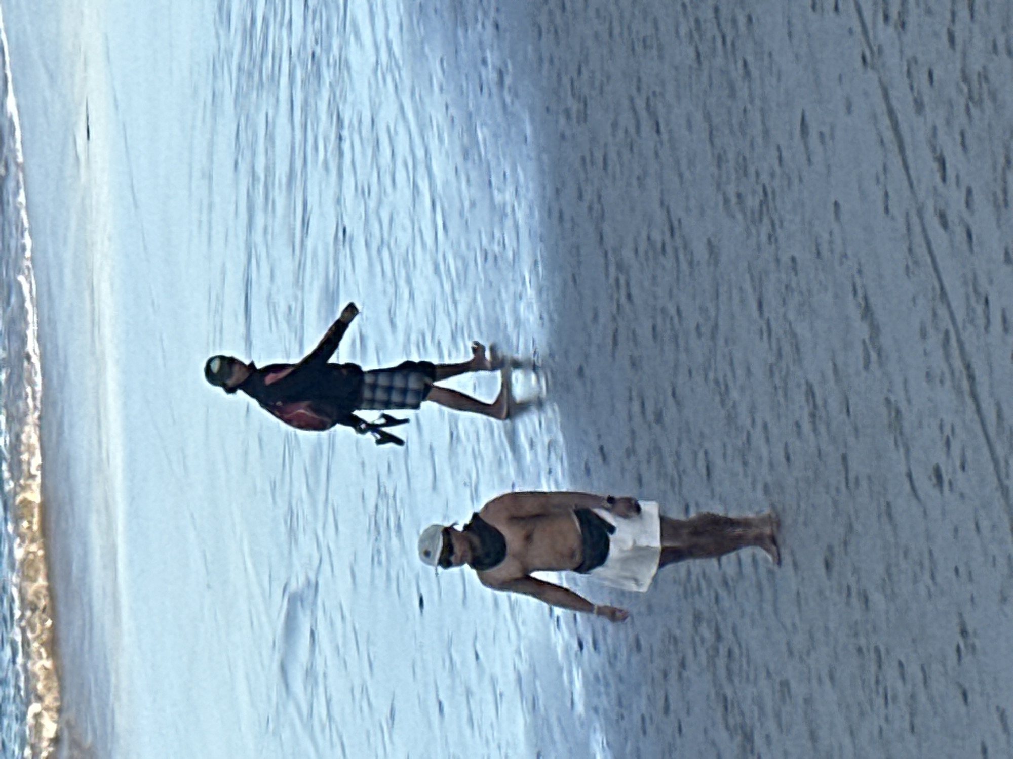 In the city of North Kuta, Indonesia, two individuals are observed on a sandy beach adjacent to a body of water. The foreground features a shirtless man wearing white shorts, a black fanny pack, sunglasses, and a light-colored baseball cap. He is facing the viewer and appears to be walking. In the mid-ground, another individual, wearing a dark jacket, plaid shorts, a backpack, and a cap, is walking away from the viewer towards the water. Both individuals are barefoot. The environment consists of a sandy beach with some scattered debris and the shallow, reflective surface of the ocean, with waves visible in the background. The lighting suggests it is daytime.