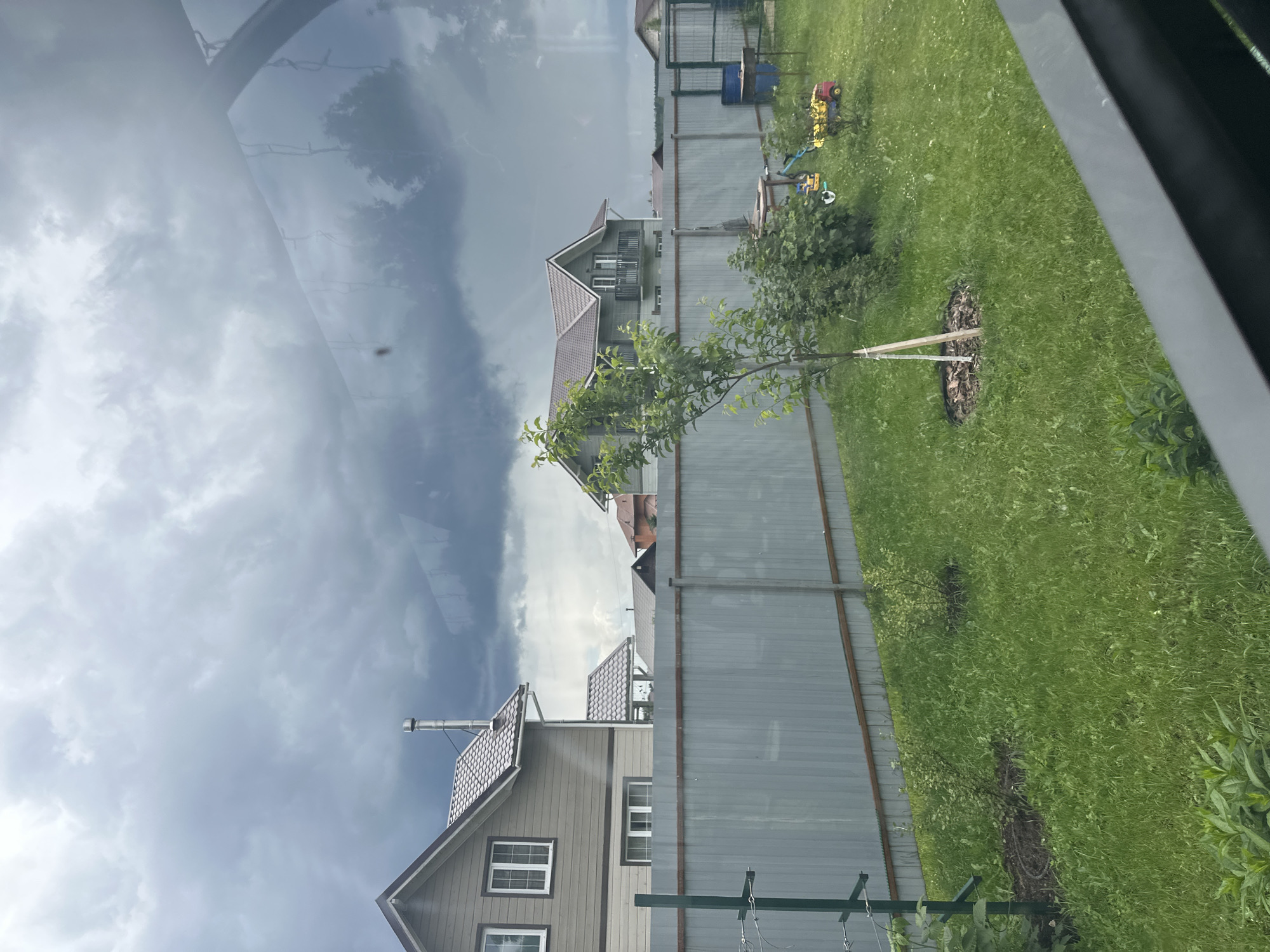 This is a vertical, outdoor photograph taken during the daytime. The scene depicts a residential area with several houses and a yard. In the foreground, a tall, grey metal fence runs horizontally across the image. Beyond the fence, a green lawn is visible, with a young tree planted in the middle, supported by wooden stakes and mulched around its base. Further back, more houses with peaked roofs and neutral-colored siding are visible. One house has a chimney and white-framed windows. The sky is overcast with dark, dramatic clouds, suggesting impending rain or a storm. Reflections are visible on a surface in the upper left of the frame, possibly a window. In the background, beyond the houses and fence, more greenery and hints of other structures can be seen. Scattered on the lawn are some children's toys, including a yellow toy truck and a blue barrel. The overall impression is of a suburban or rural setting on a cloudy day.