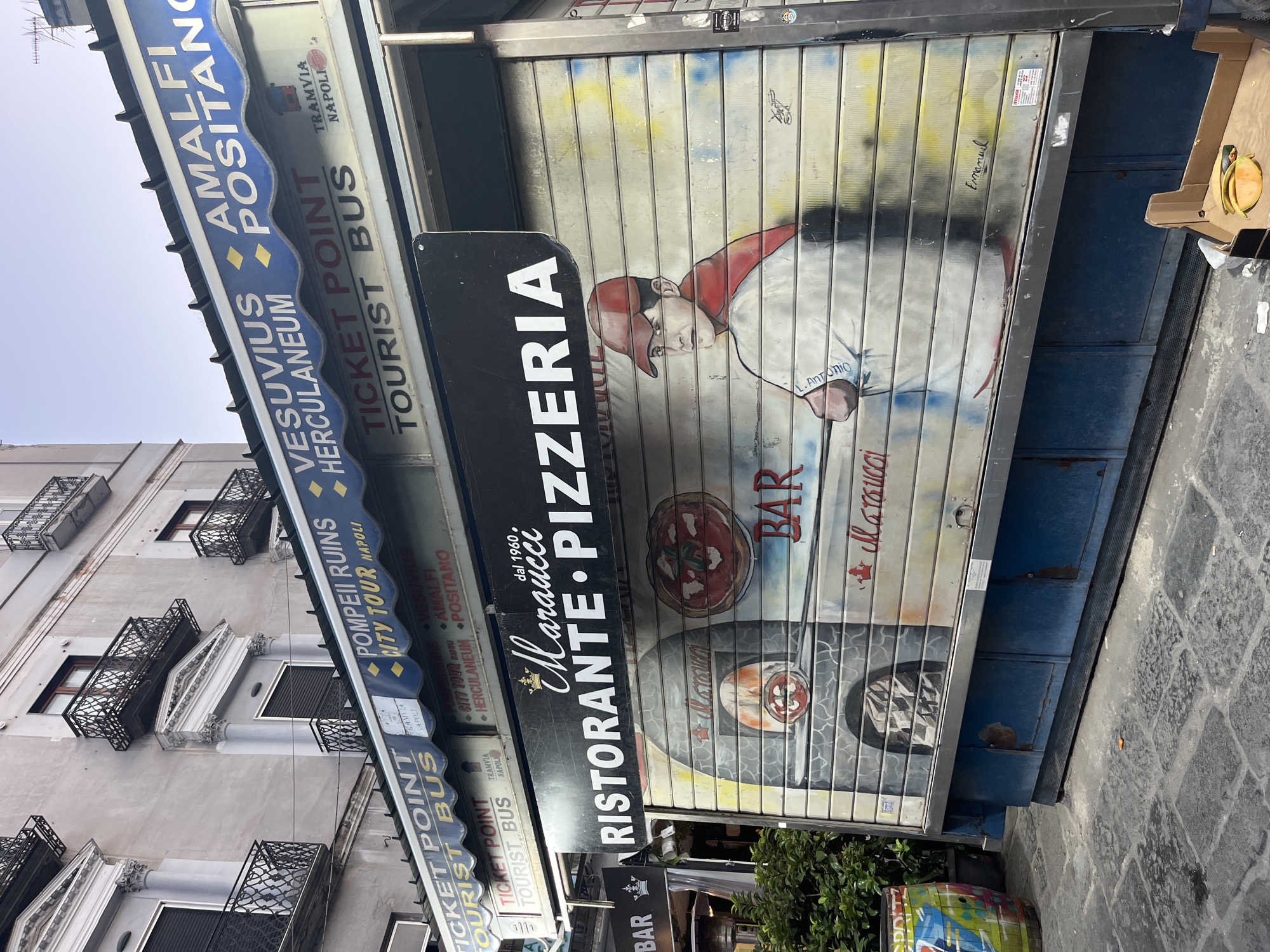 The image depicts a street-level view of a closed pizza restaurant and ticket sales booth in Naples, Italy. The establishment, named "Maraucci Pizzeria," features a large, rolled-down metal shutter adorned with a mural. The mural displays a pizza oven with a pizza inside, and a young man wearing a red cap and a red scarf, looking towards the viewer. The text "Ristorante Pizzeria dal 1960" is prominently displayed above the mural.
Above the shuttered pizza place, a blue awning displays advertising for various tourist attractions and transportation. Text on the awning includes "TICKET POINT TOURIST BUS," "VESUVIUS HERCULANEUM," "AMALFI POSITANO," and "POMPEII RUINS CITY TOUR NAPOLI."
To the left of the pizza establishment, a smaller sign for "BAR" is visible, with an outdoor heater and some greenery behind it. The street in front of the businesses is paved with stone cobbles. In the lower right corner, a cardboard box contains cut fruit, possibly melon and cucumber, and there is some litter on the ground. The building behind the awning has a classical architectural style with balconies. The sky is overcast.