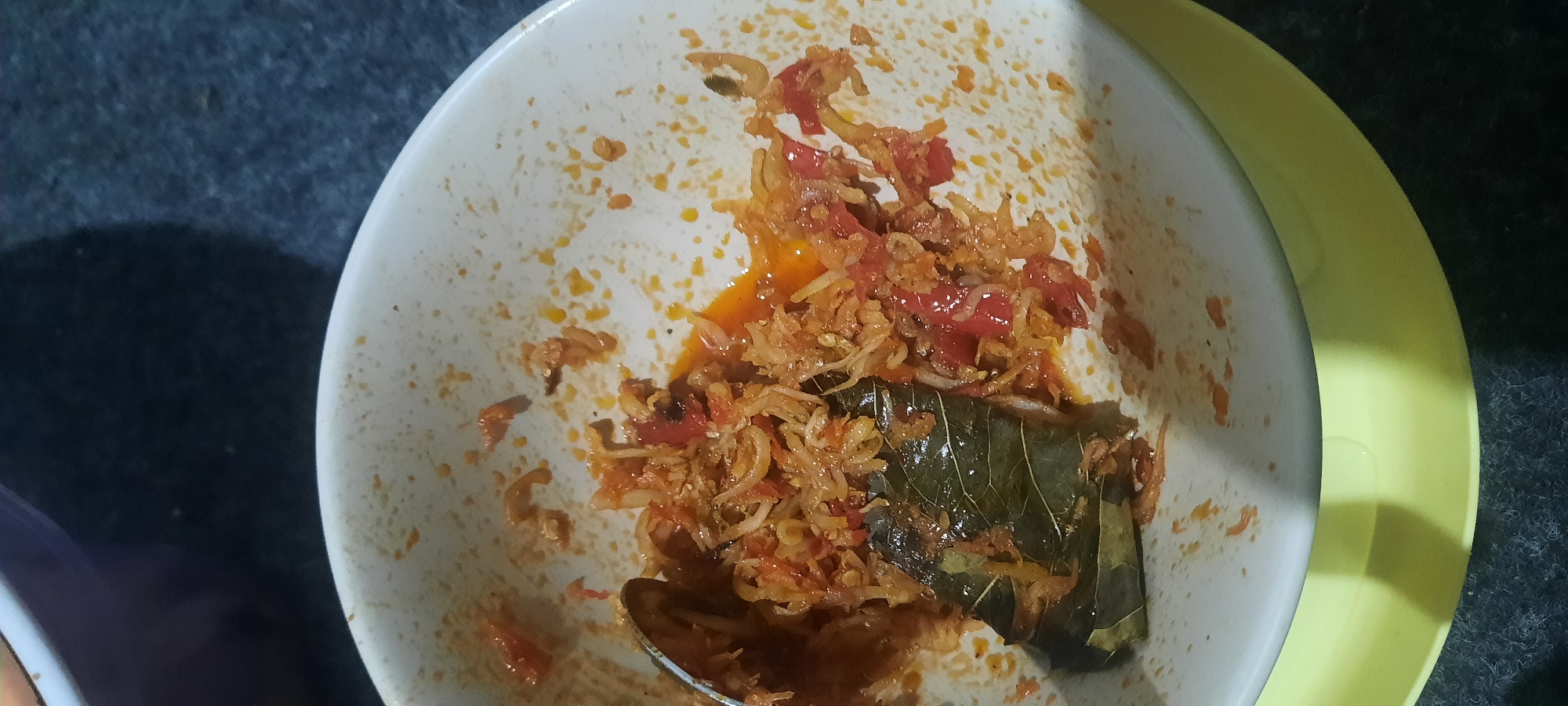 A close-up, overhead shot shows a white bowl containing a red, soupy mixture of what appears to be chopped tomatoes, shallots, and small fish, possibly anchovies. A single, large green leaf is partially submerged in the mixture, and a metal spoon rests on the left side of the bowl. The bowl sits on a yellow plate, which is placed on a dark, textured surface, likely a table. The background is dark and out of focus. The scene is indoors, and the lighting is dim, suggesting it may be evening or night. There are no people visible in the image. The image appears to be taken in North Jakarta, Indonesia, where this type of dish, potentially a spicy fish and tomato relish called "sambal," is commonly consumed.