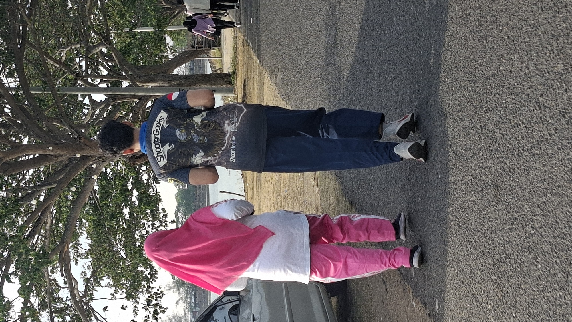 Two individuals are walking away from the camera on an asphalt path next to a body of water. The person on the left is wearing a bright pink hijab and pink pants with white stripes, and a white long-sleeved shirt. The person on the right is wearing a dark t-shirt with a graphic of an eagle and the text "Shooter Geeks" and blue pants. The path is lined with trees, and in the background, other pedestrians are visible walking in the same direction. A silver car is parked on the left side of the frame. The scene appears to be in a park or recreational area in Lhokseumawe, Indonesia, possibly near the Ujong Blang beach area given the presence of trees and water, and the general ambiance. The lighting suggests it is late afternoon or early morning.