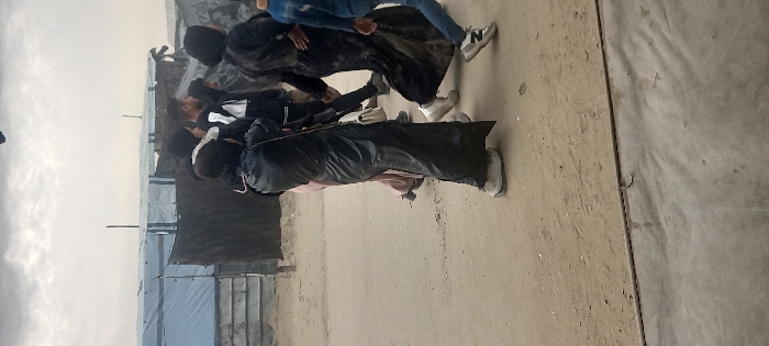 The image depicts a group of individuals walking across a dirt path in what appears to be a makeshift settlement within Khan Yunis, Palestinian Territory. The ground consists primarily of light brown compacted earth, transitioning to a strip of gray concrete or pavement at the bottom of the frame.
In the foreground, a person is walking from left to right, wearing a black, full-length garment that covers the head, and light-colored athletic shoes. A white shoulder bag with a gold chain strap is visible across their body. Behind this individual, another person in a light pink or peach full-length garment is partially visible. Further into the midground, several other individuals are walking in the same direction. One person wears a dark, possibly black, head covering and long garment, paired with white athletic shoes. Another person, identified as male, has short dark hair and wears a dark jacket with white accents and dark pants. Various other figures, mostly obscured, are also present, generally wearing dark clothing. To the far right, the lower half of a person is visible, wearing blue jeans, a blue top, and two-toned athletic shoes.
The background features multiple temporary structures constructed from various materials, including blue and white tarpaulins and dark fabric, indicative of tents or shelters. Vertical wooden or metal poles support some of these structures. The sky overhead is overcast, displaying a uniform light gray cloud cover. The overall scene suggests movement within a populated but informally constructed area.