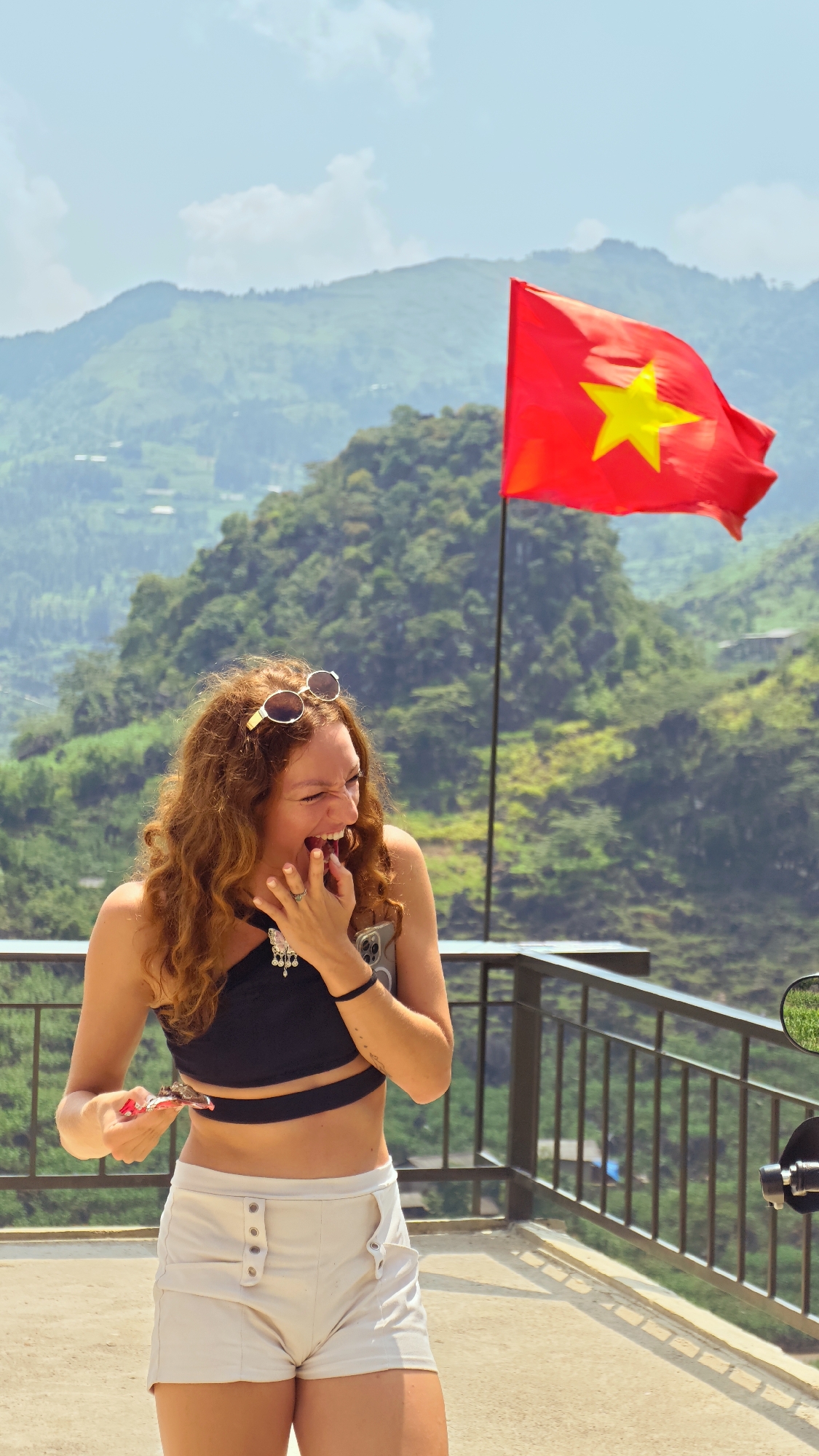 A woman with curly red hair is laughing with her hand over her mouth on a balcony. She is wearing a black crop top, white shorts and sunglasses on her head. She is holding a small object in her other hand. In the background there is a Vietnamese flag waving in the wind against a backdrop of green mountains. There is a metal railing separating the balcony from the mountains. The scene is set in Lũng Phìn Commune, Vietnam.