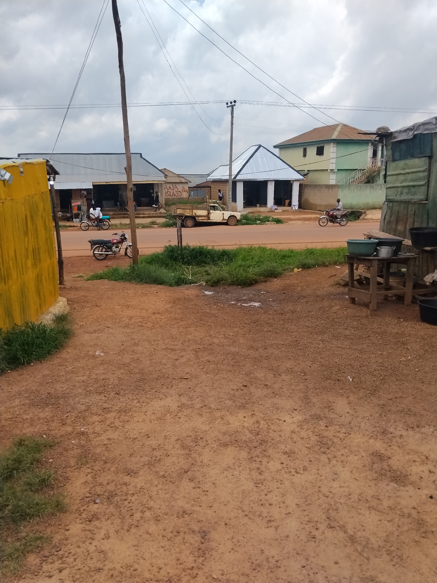 The image depicts a street scene in Durumi, Nigeria, with a focus on a dusty road and buildings lining it. In the foreground, a patch of dry dirt ground is visible, with sparse green grass growing on the left side. A tall wooden pole with overhead wires stands prominently in the middle of the frame.
To the left of the pole, a bright yellow corrugated metal structure is partially visible. Across the road, a row of shops with corrugated metal roofs are situated. One shop has text that reads "AJA ISLAND B" painted on its facade. A light-colored pickup truck is parked in front of one of the shops.
Several motorcycles are present: a red motorcycle is parked on the right side of the road, and another motorcycle with a rider is seen further down the road. A person is also visible riding a motorcycle in the middle of the road.
In the background, a two-story building with green and yellow walls and a brown roof is visible. To the right of this building, another structure with green corrugated metal walls is present. On a wooden table in front of the green structure, several dark basins and a metal pot are displayed. The sky is overcast with clouds.