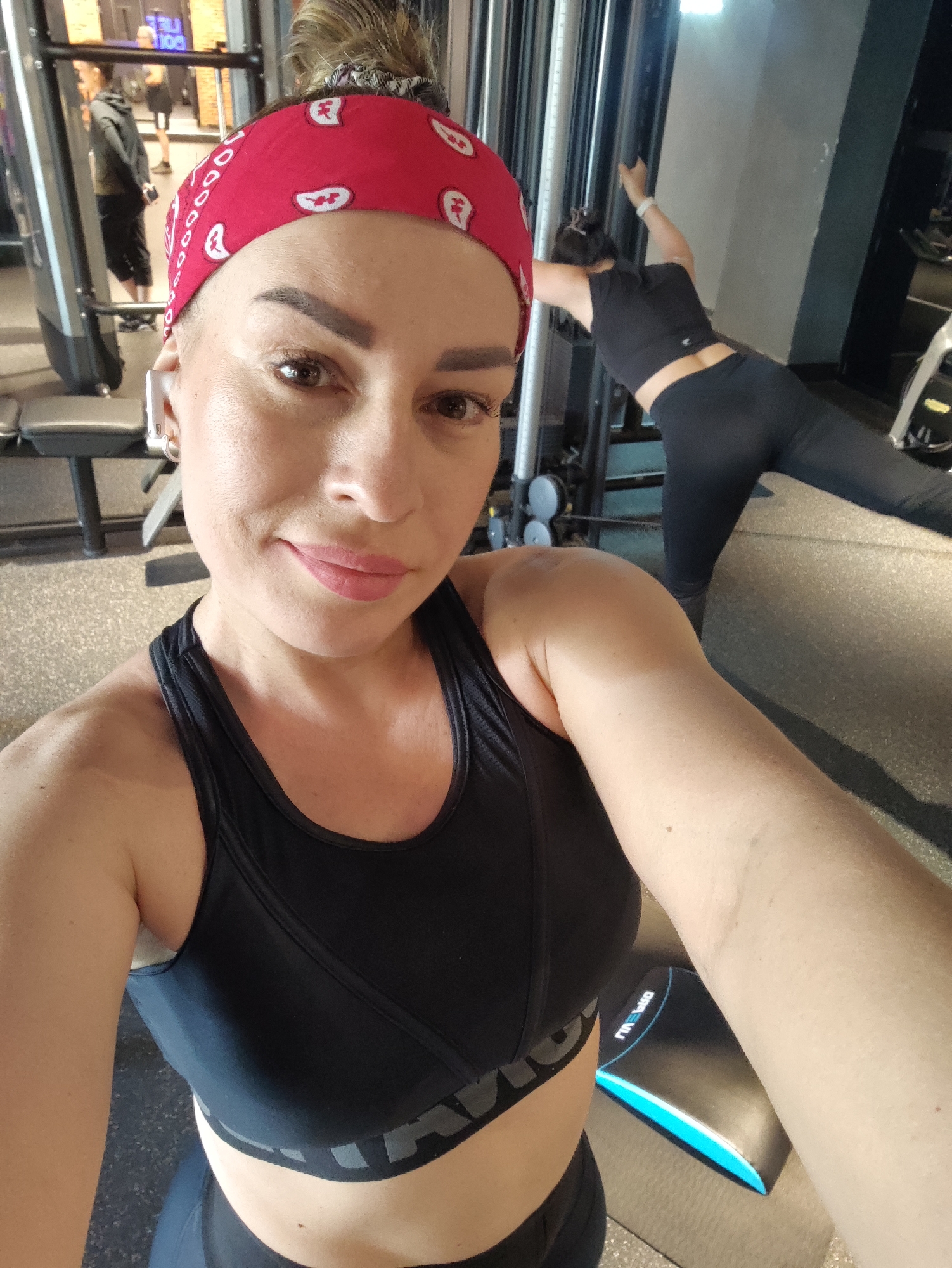 A woman in a black sports bra and a red bandana is taking a selfie in a gym. She is wearing white wireless earbuds. In the background, another woman is exercising. The gym appears to be modern, with weight machines and grey walls. The flooring is a dark speckled material. The image was taken in Almaty, Kazakhstan.