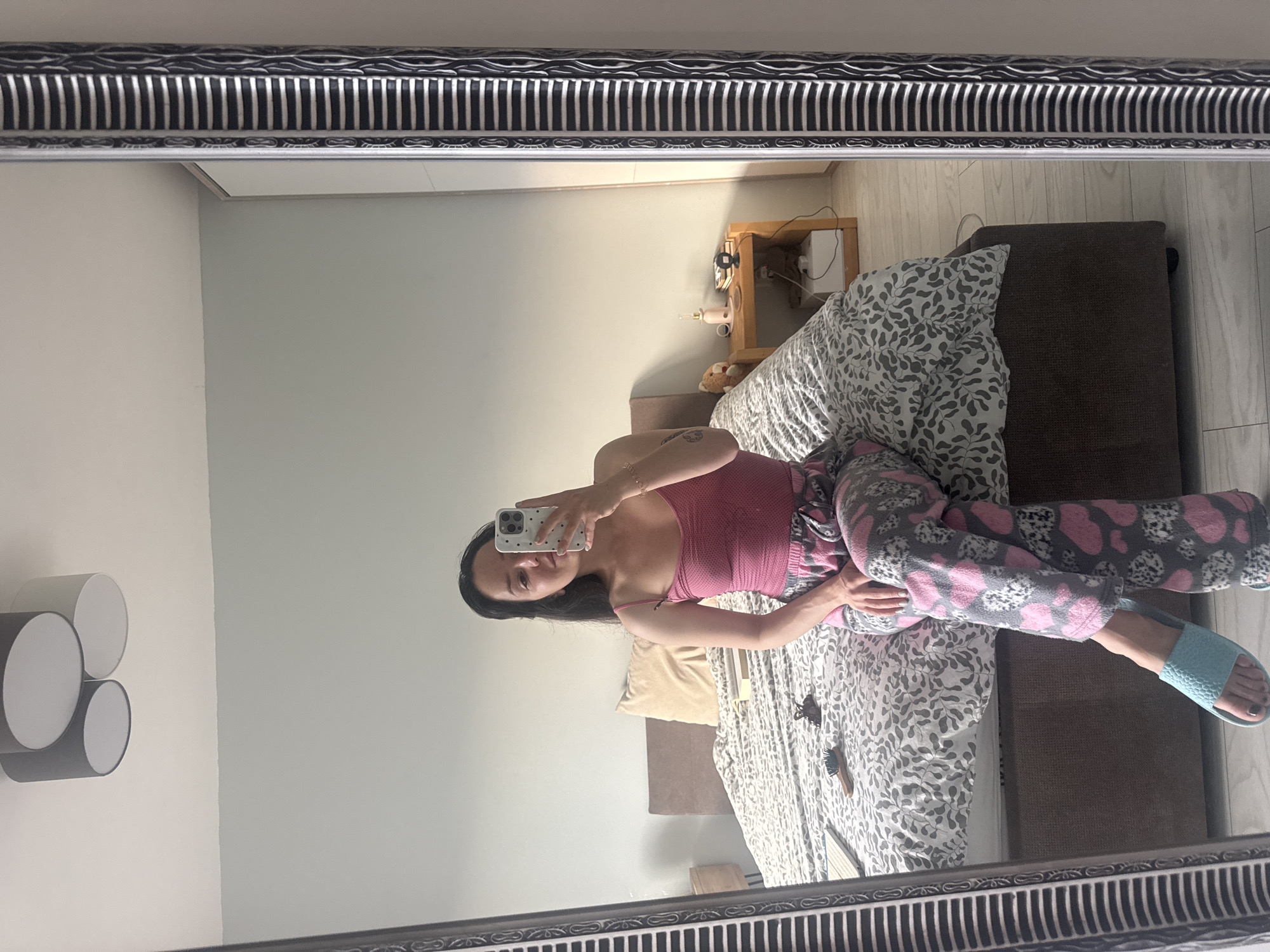The image is a mirror selfie showing a woman reclining on a bed in a bedroom. The woman, an adult with long dark hair, is wearing a pink tank top and grey pajama bottoms patterned with pink hearts and white/grey leopard print spots. She also has light blue open-toed slippers on her feet. She holds a white smartphone with a black spotted case in her right hand, using it to capture her reflection in the mirror. A tattoo is visible on her upper left arm, and a bracelet adorns her left wrist. Her expression is neutral as she looks towards the phone.
The mirror she is using has a distinctive silver-colored, ornate frame featuring vertical ridged patterns and wavy textures. The bed has a white duvet cover with a grey leaf or floral pattern and a light yellow pillow. The bed frame appears to be upholstered in a dark brown or grey fabric. Next to the bed, a light wooden nightstand holds a small pink lamp-like object and some electrical cords. A small teddy bear is partially visible on the floor or next to the bed.
On the wall to the left, there is a modern wall light fixture comprising four cylindrical shapes, two in white and two in dark grey. A brown hair clip and a small wooden-handled brush are lying on the bed near the woman's legs. The walls are light-colored, and the floor visible in the background is light wood or laminate. The scene depicts a casual, relaxed moment in a home setting, consistent with a bedroom in a private residence, potentially in Burgas, Bulgaria. No text is visible in the image.