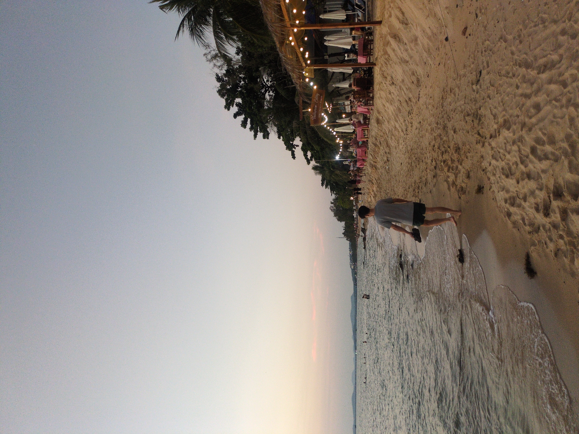 The image shows a beach scene at sunset in Phú Quốc, Vietnam. A man in a gray shirt and dark shorts walks along the shoreline, holding his shoes in his hand. The beach is sandy, and gentle waves are lapping at the shore. Along the edge of the beach is a restaurant or bar area with thatched roofs, string lights, and tables with pink tablecloths. Palm trees are visible along the coastline. The sky is a pale blue-gray, indicating the early evening light.