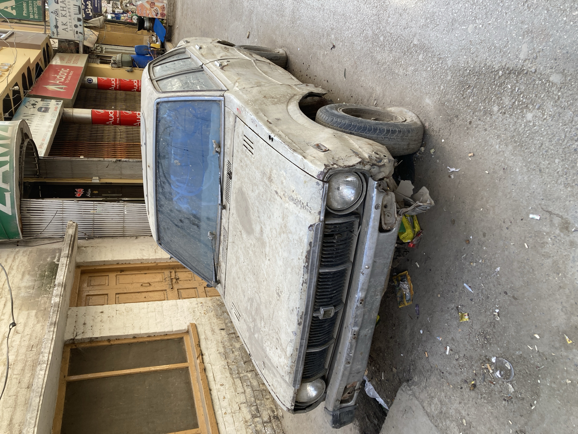 The image depicts a severely damaged, dilapidated car parked on a street in Quetta City Tehsil, Pakistan. The car appears to be an older model, heavily soiled, with visible damage to the front end and missing components. The front tire is flat. Litter and debris surround the vehicle on the pavement. Behind the car, there is a row of buildings with closed metal shutters on shopfronts and various signs, including "AK KHA ENTERPRISES", "Audionic" and "HIKVISION". The buildings have a faded appearance with visible wear and tear. The general environment appears to be an urban commercial area in a state of disrepair.